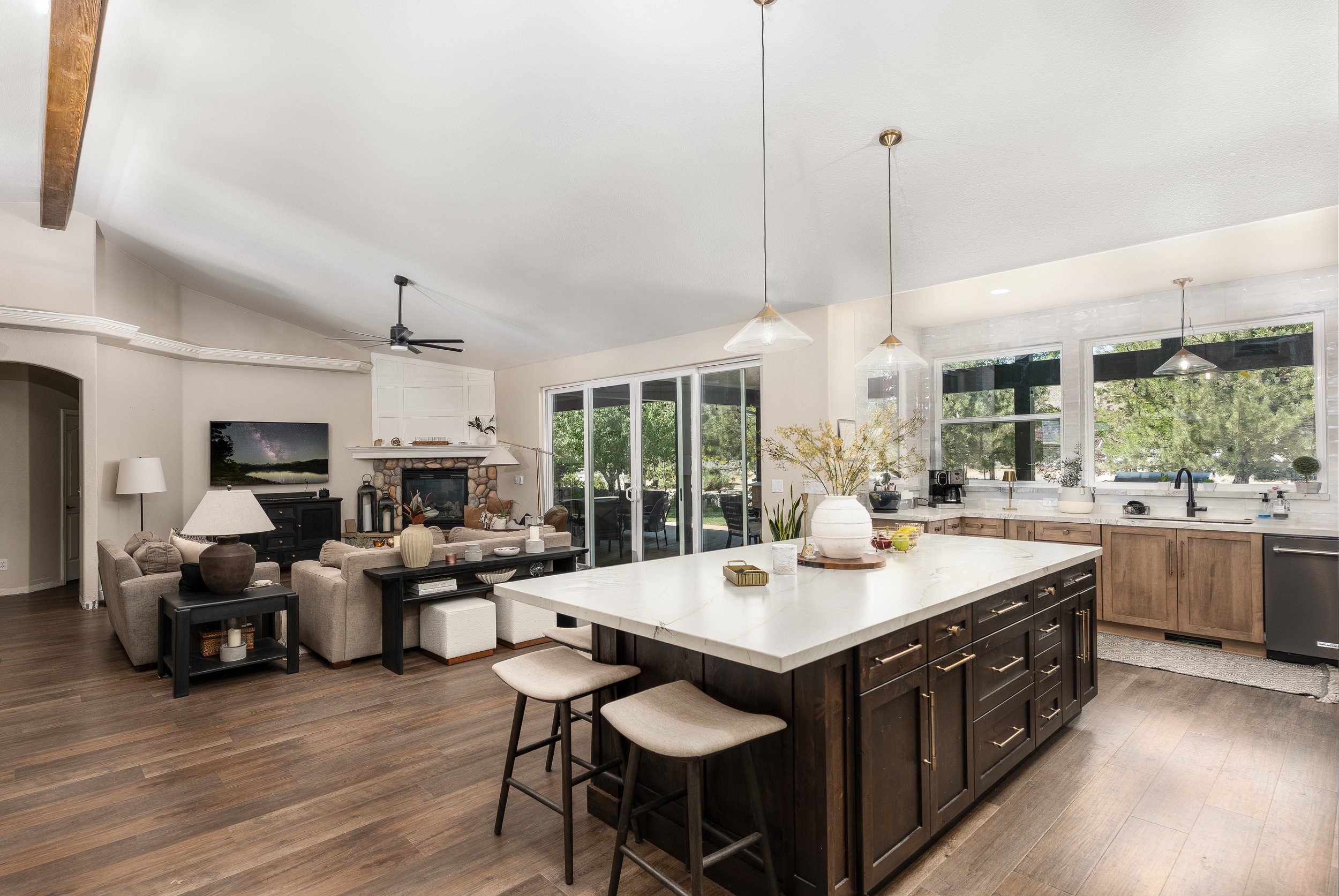 Open-concept living room and kitchen with white walls, wood flooring, large windows, kitchen island with barstools, and a seating area with sofas and a TV.