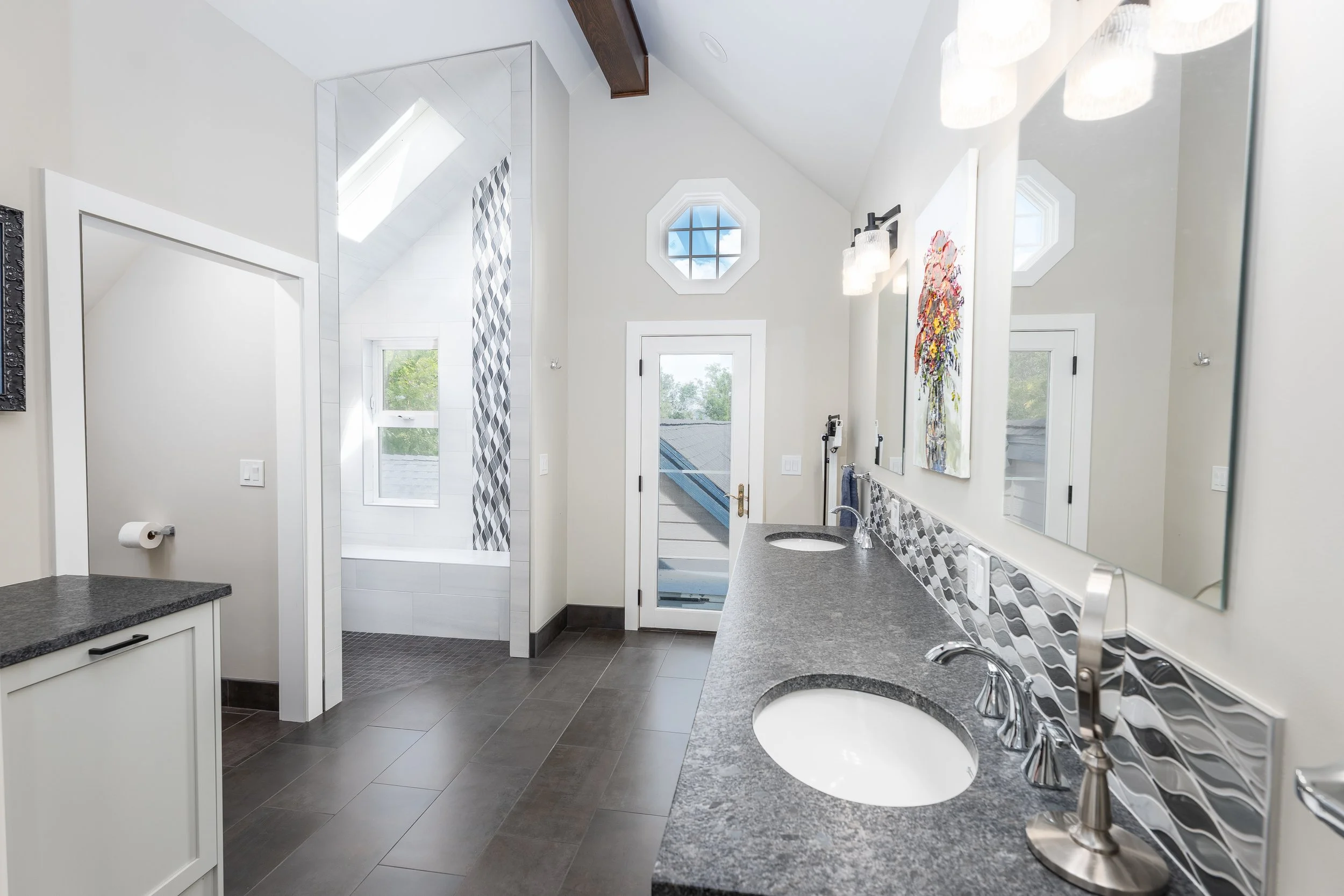 Modern bathroom with double sink granite countertop, large mirror, patterned backsplash, art on wall, and a door leading outside, with a bathtub visible through an open doorway.