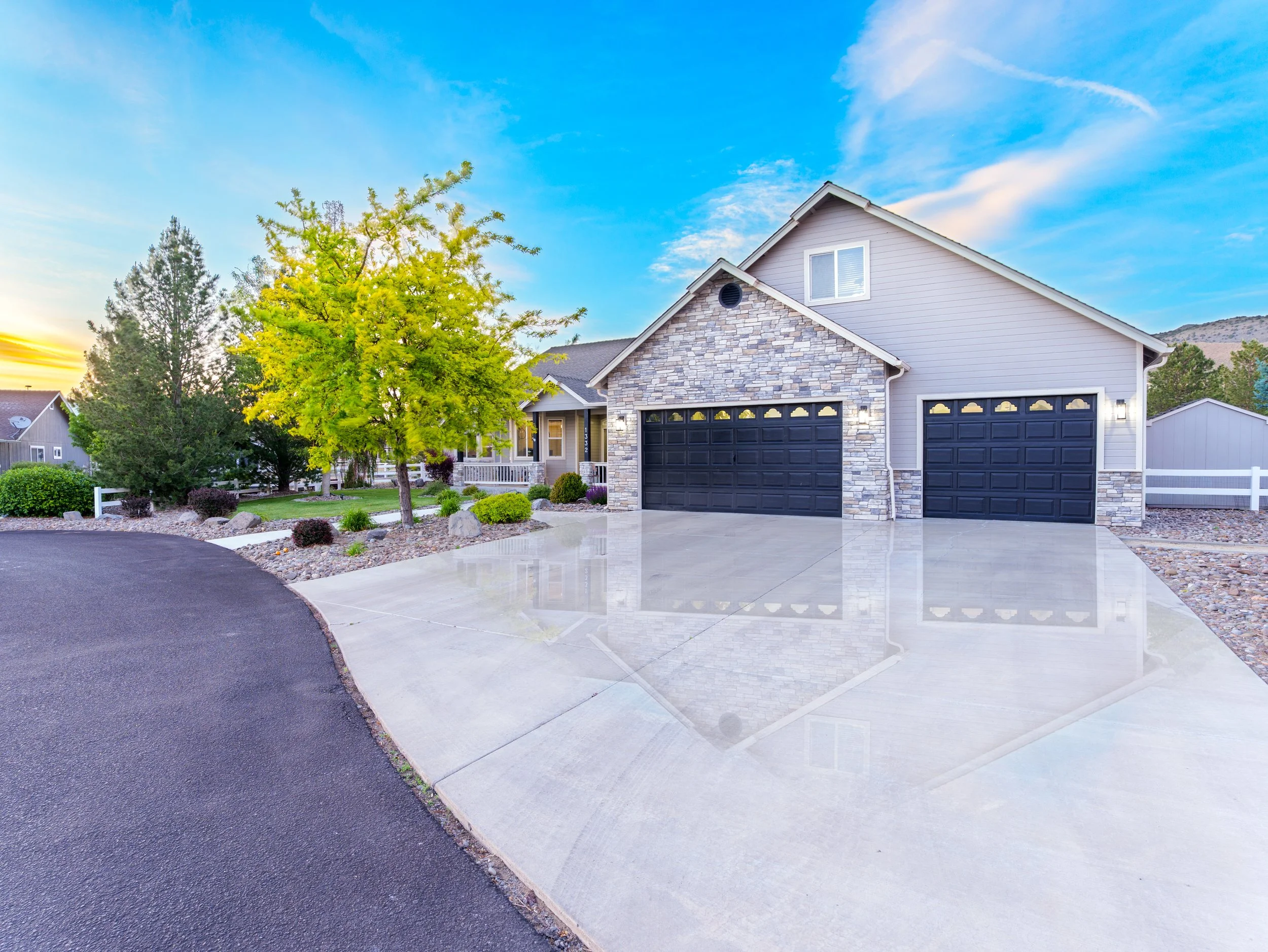 Suburban house with a grey exterior, stone accents, and a black garage door, surrounded by landscaping and trees with a blue sky in the background.