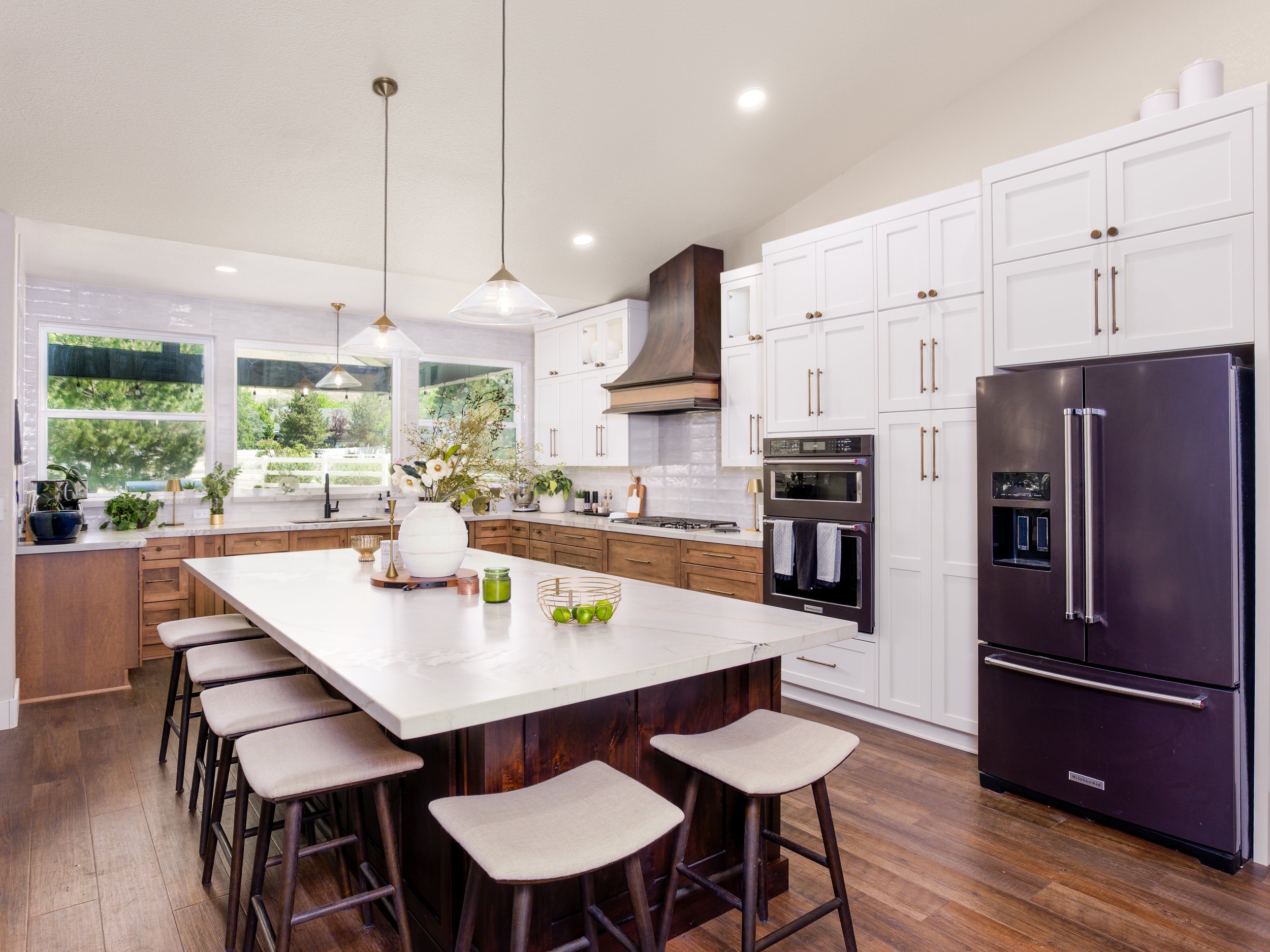 Modern kitchen with white cabinetry, a large central island with chairs, wood flooring, a stainless steel refrigerator, and large windows with a view of greenery outside.
