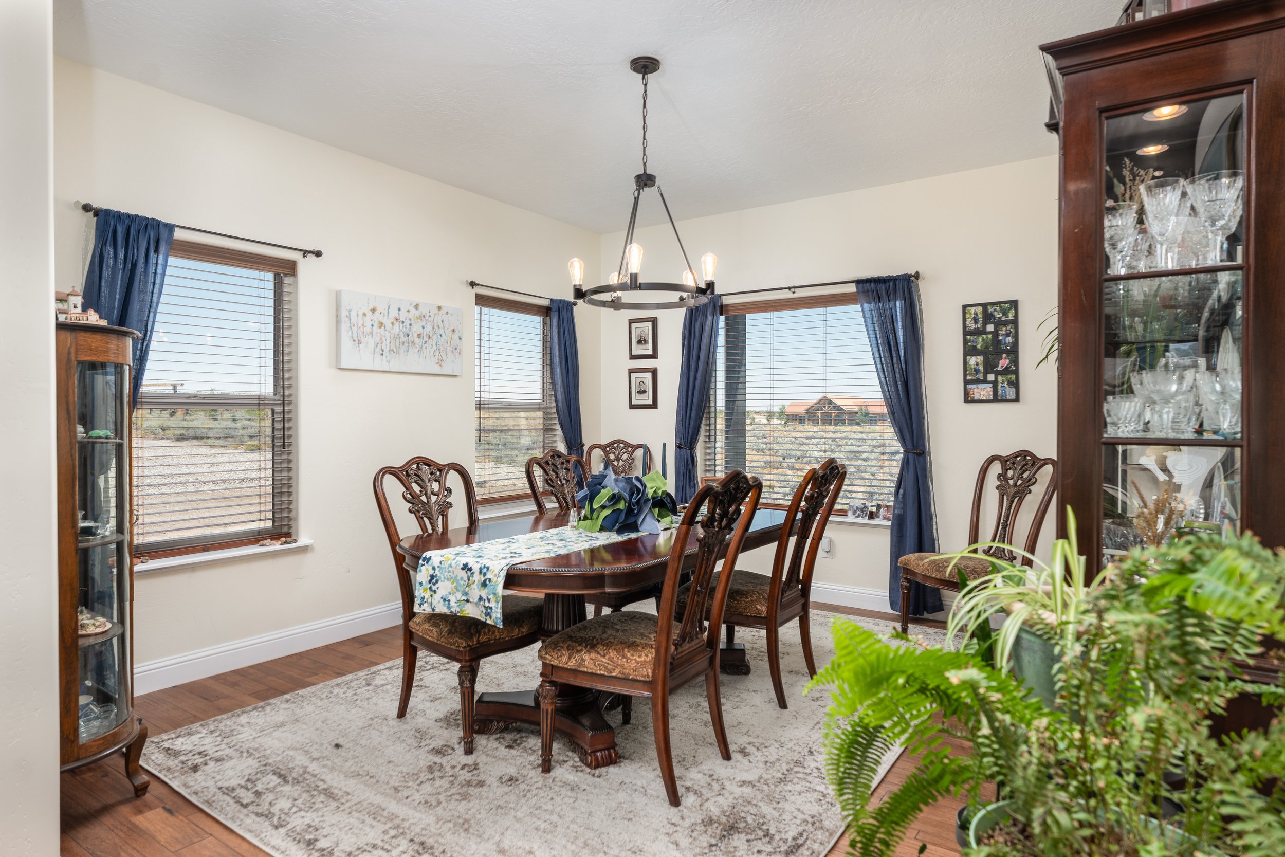 A dining room with a wooden table and six carved wooden chairs, a beige rug, a china cabinet with glassware, and plants, with three windows with blue curtains and a modern chandelier hanging from the ceiling.
