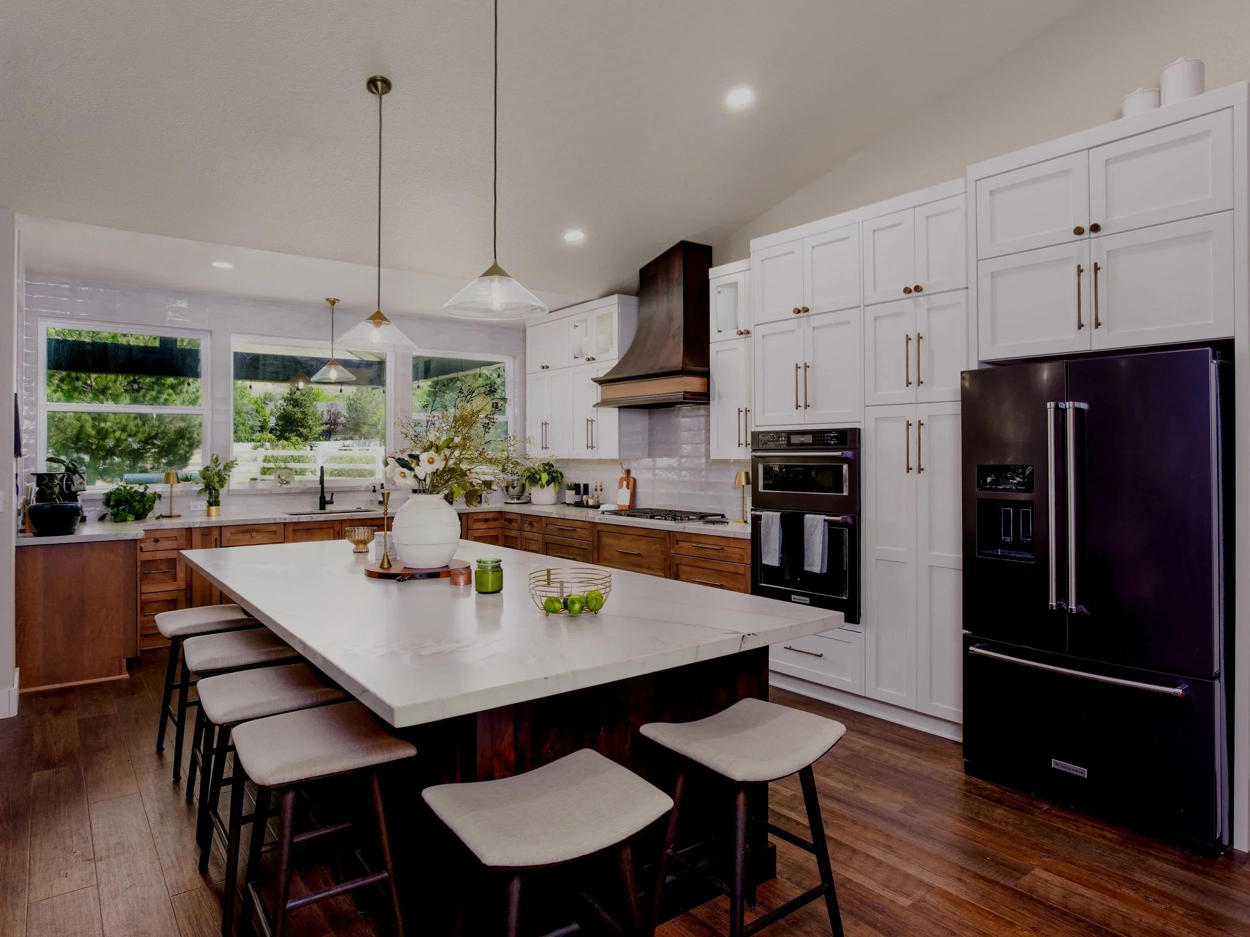 Modern kitchen with white cabinets, wooden lower cabinets, black appliances, and large windows bringing in natural light; decorated with plants and flowers.