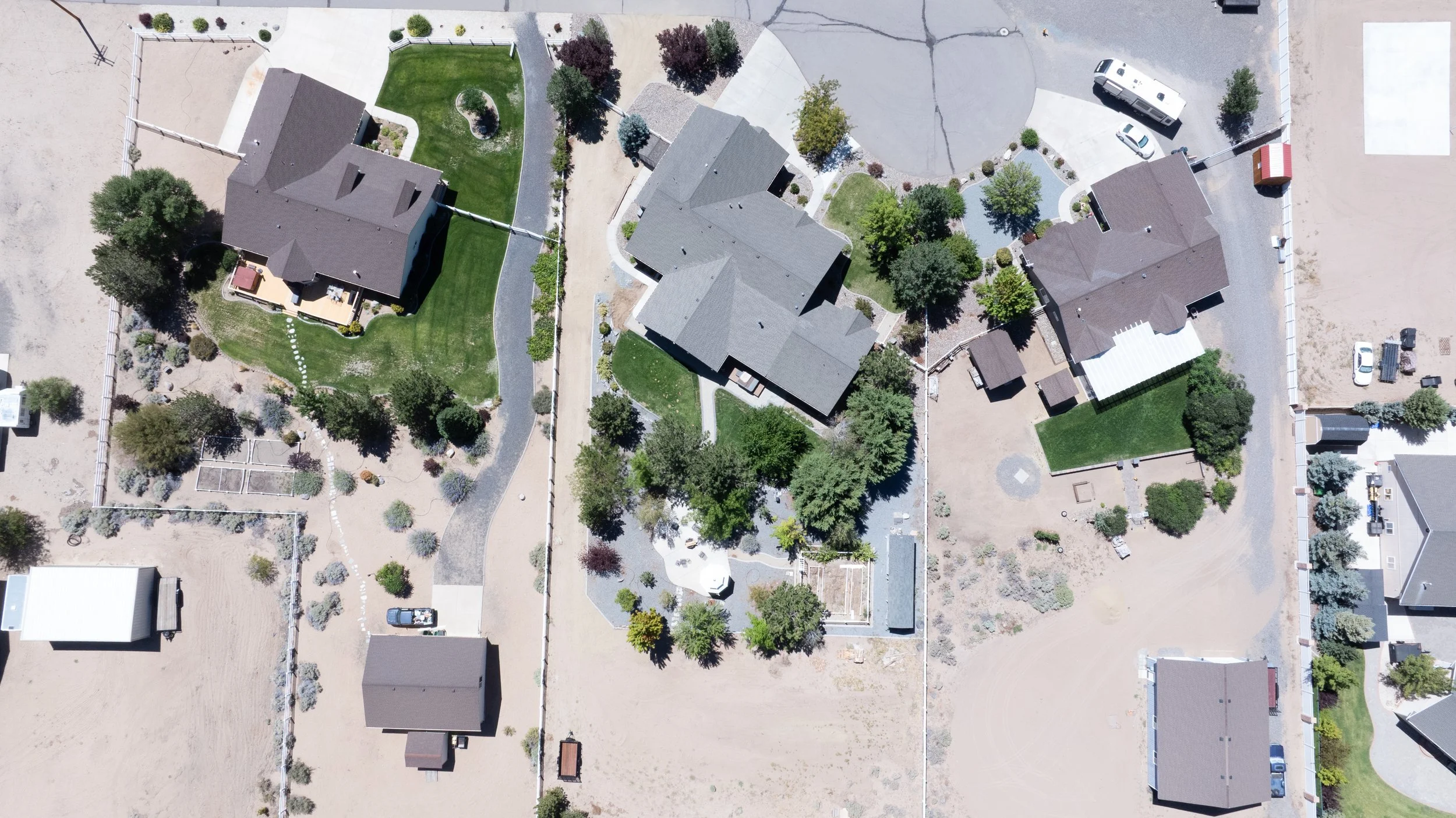 Aerial view of a residential neighborhood with several houses, trees, lawns, driveways, and some parked vehicles, with a mix of green and barren areas.