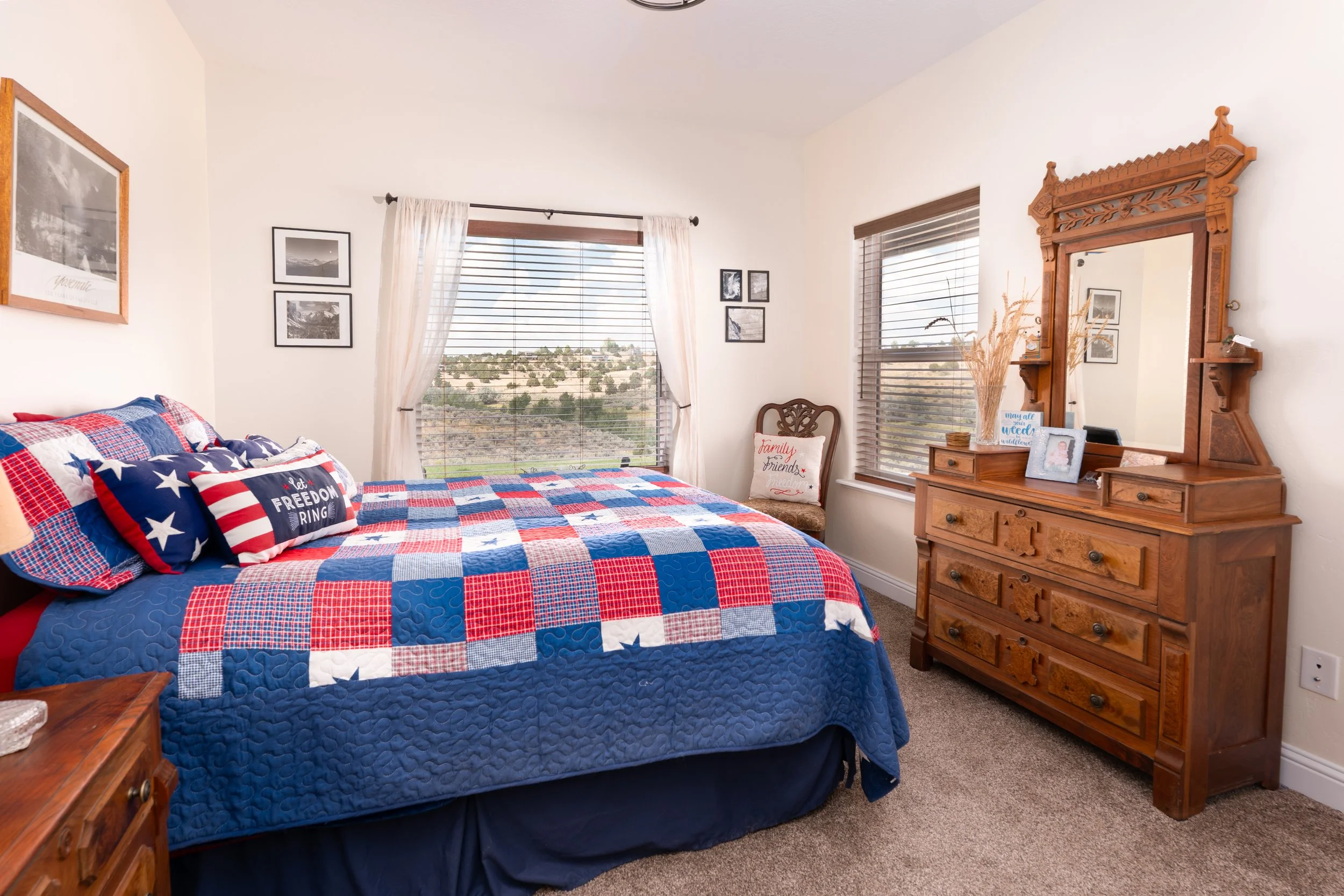 A bedroom decorated in patriotic colors with a bed that has red, white, and blue quilt, and pillows featuring stars and stripes. There is a wooden dresser with a mirror, a brown chair with a pillow, and windows with blinds and curtains showing a hill