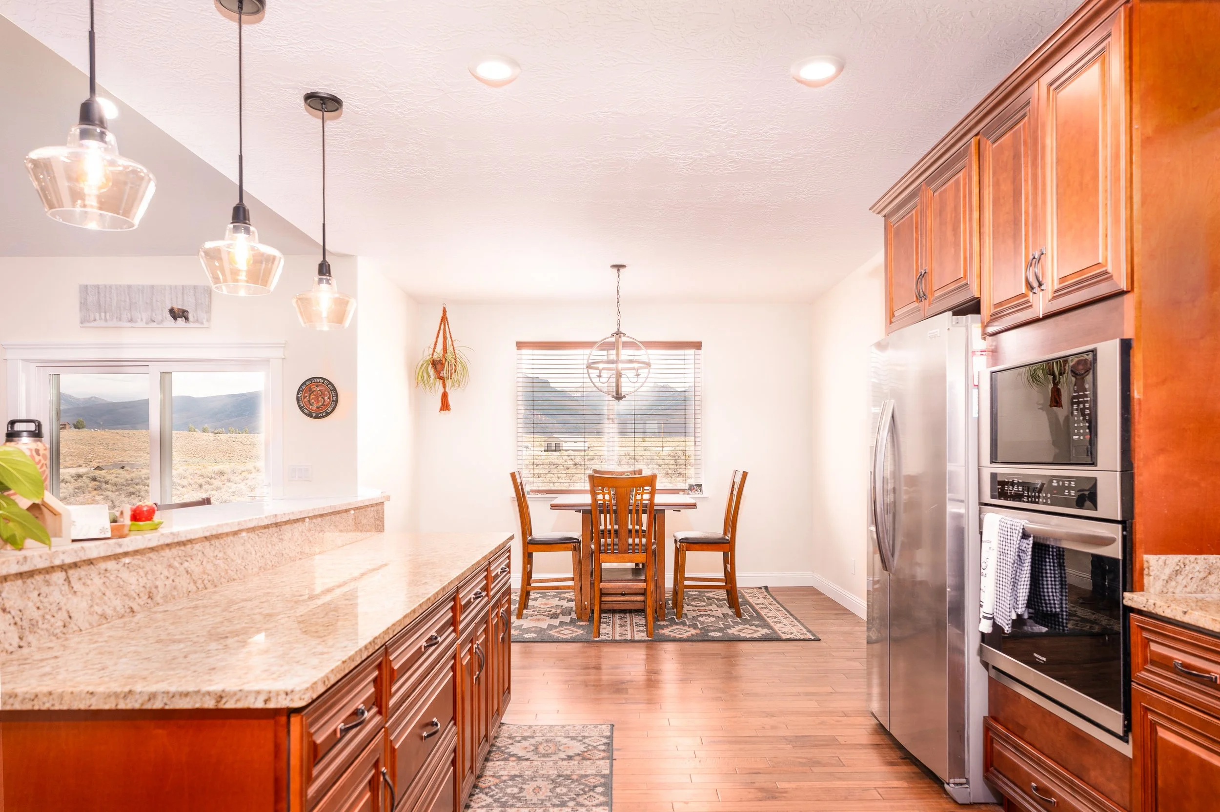 Bright kitchen and dining area with wooden cabinets, granite countertops, hardwood floors, and a window overlooking a mountainous landscape.