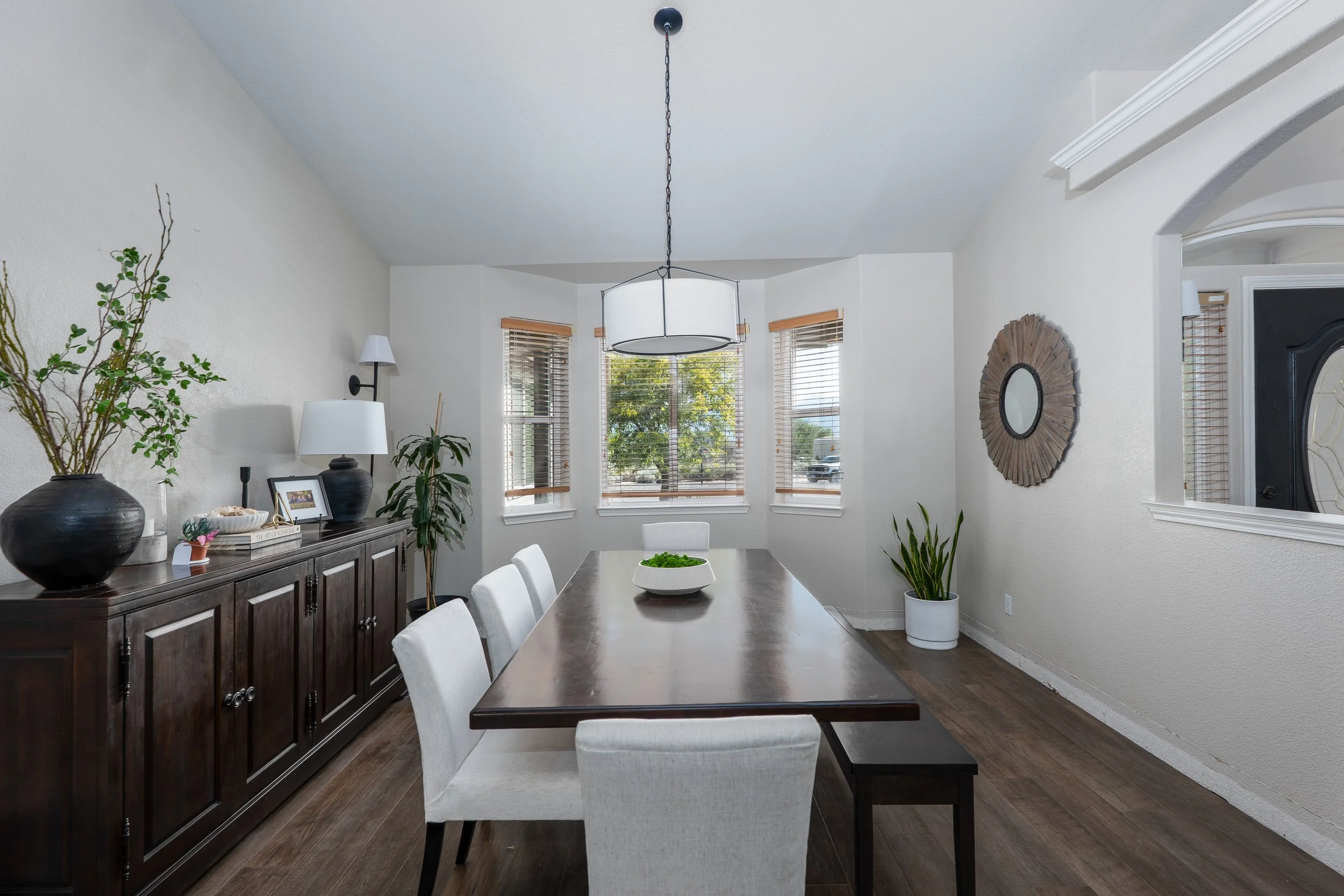 A dining room with a wooden table, six upholstered chairs, a dark sideboard with decorative items, plants, and a window with blinds letting in natural light.