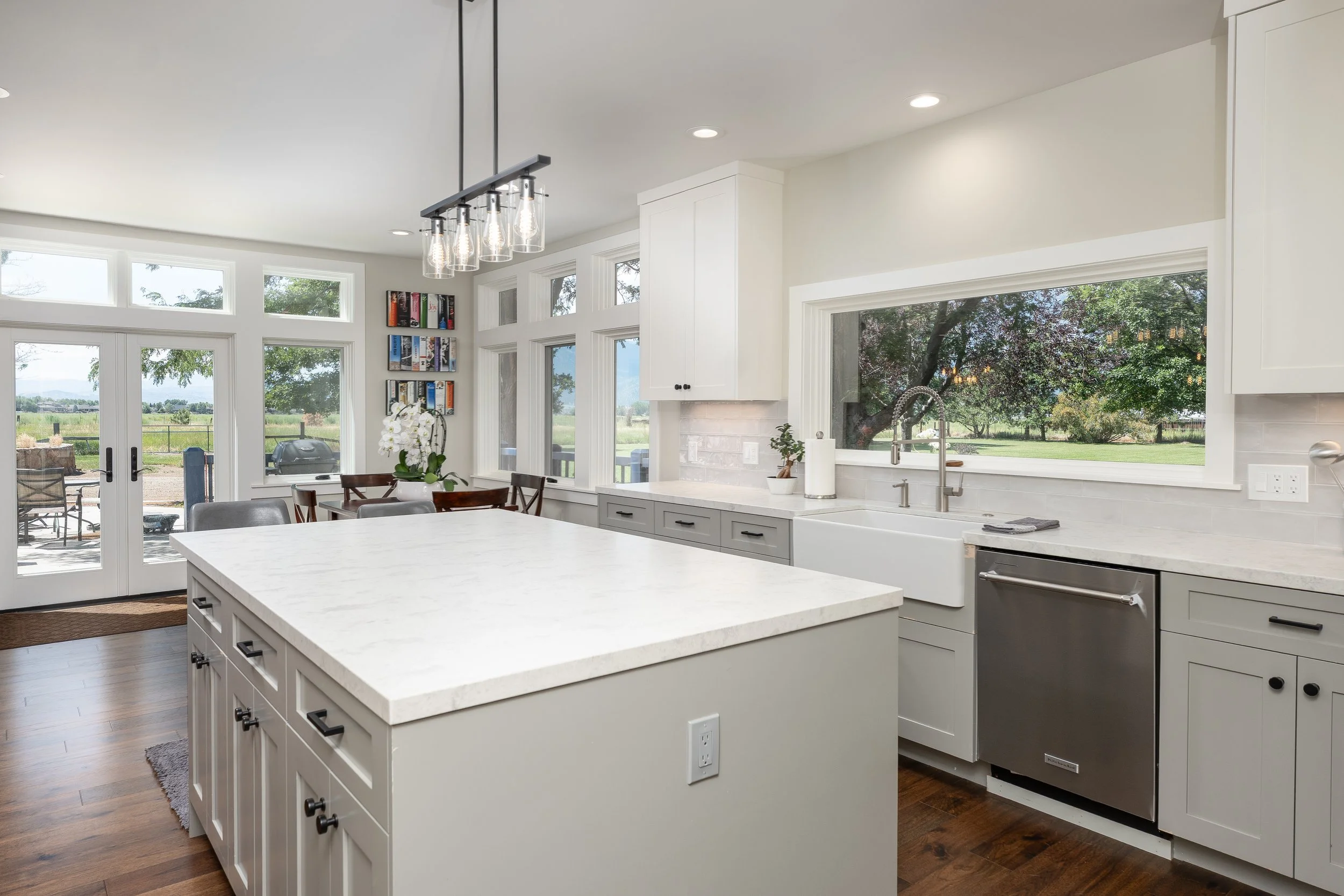 Modern kitchen with white cabinets, a large island with a white countertop, large windows and glass doors showing a backyard with trees and a lawn, and a black chandelier with hanging Edison bulbs.