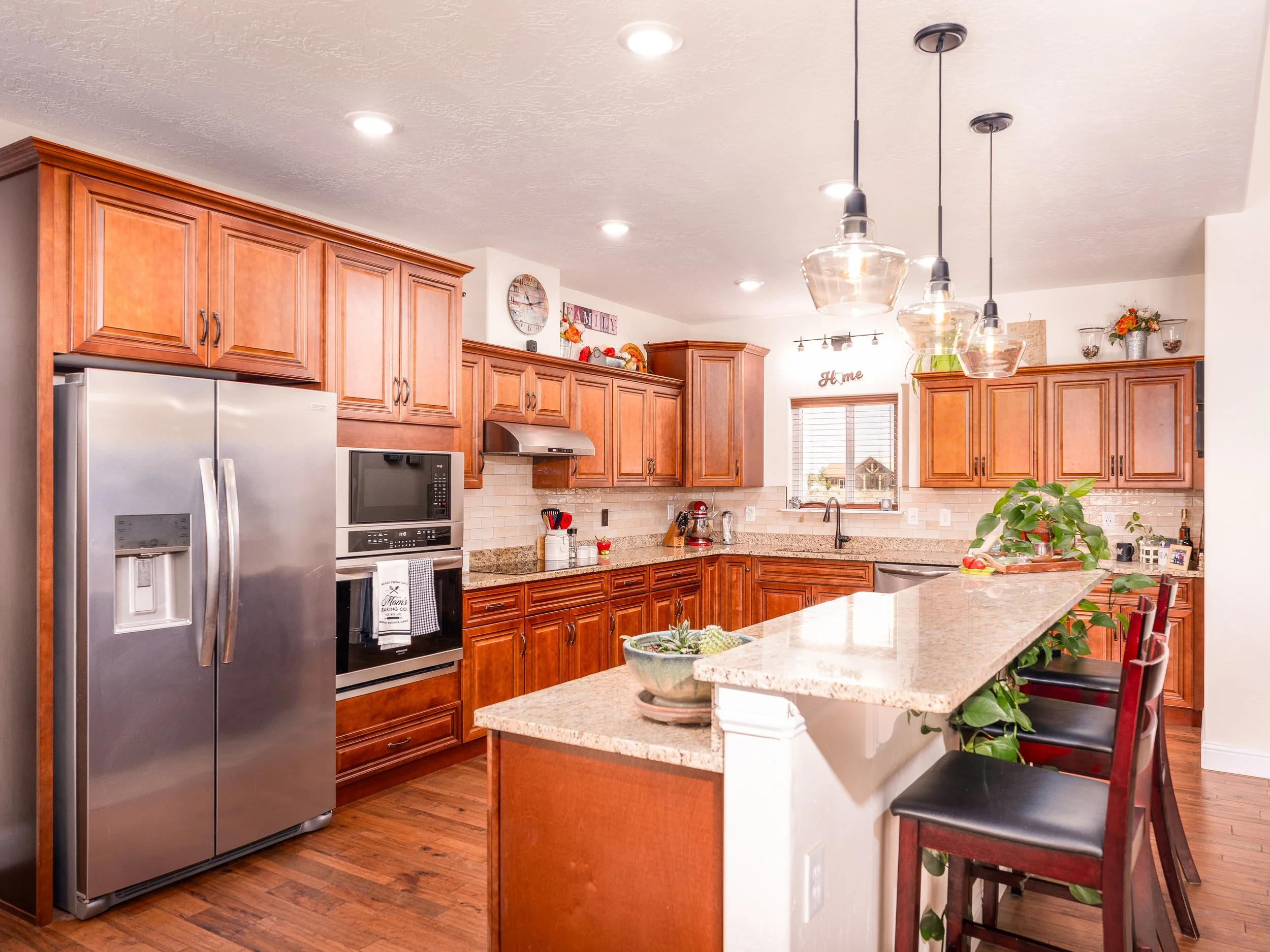 A spacious kitchen featuring wooden cabinets, stainless steel appliances, a granite countertop, and decorative elements such as plants, a clock, and wall signs.