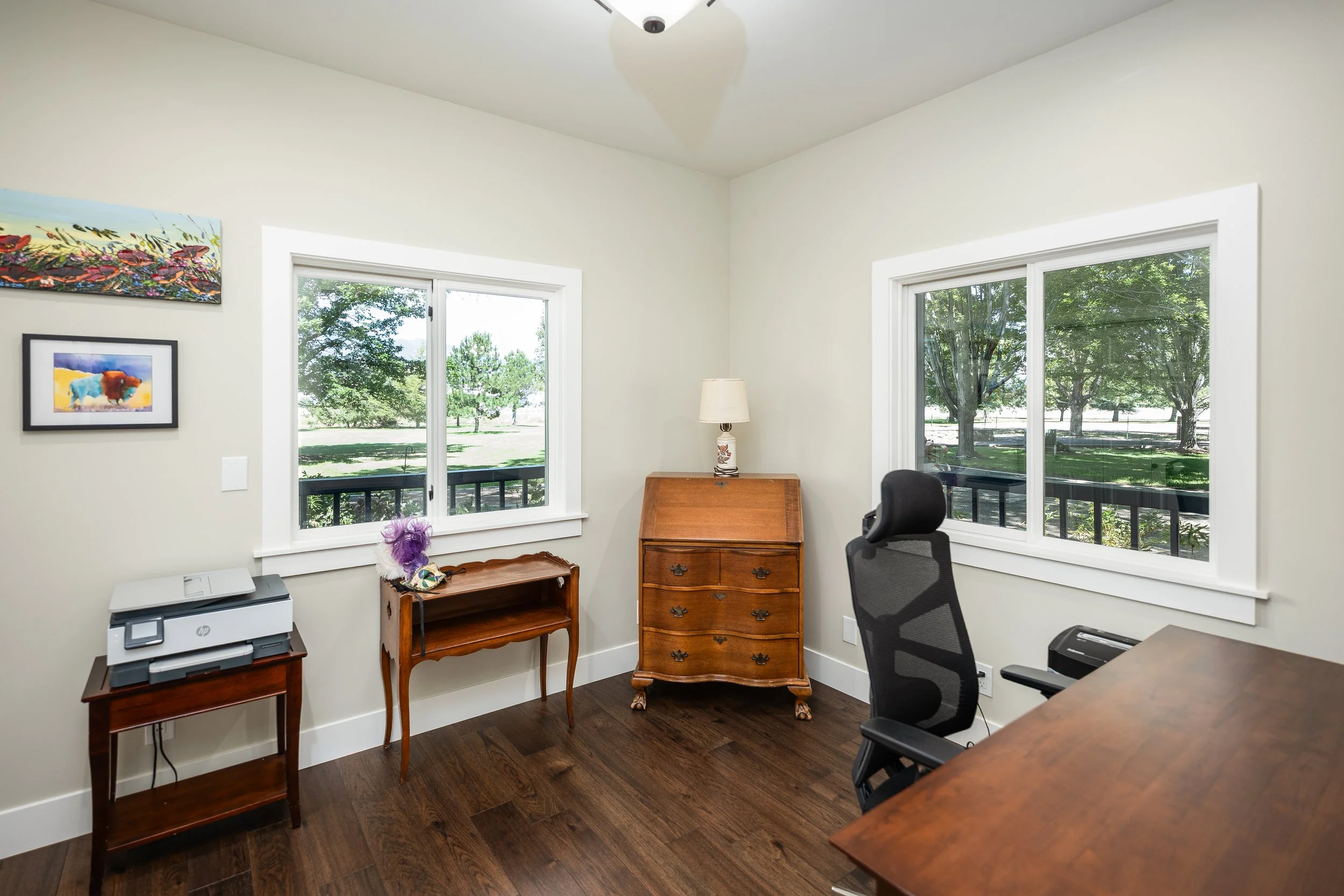 Home office with two windows, wooden furniture, a black office chair, and art on the wall.