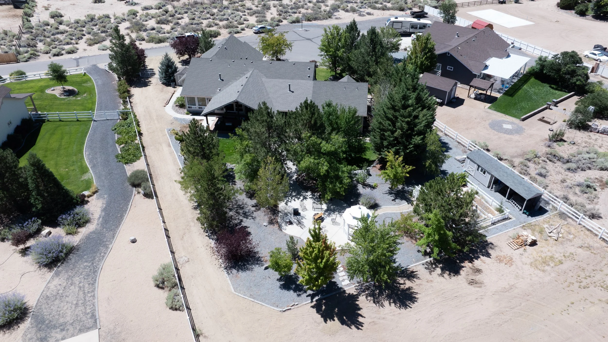 Aerial view of a residential backyard with a large house, multiple trees, a circular patio, and surrounding fencing. Adjacent desert landscape with sparse plants and dirt areas.