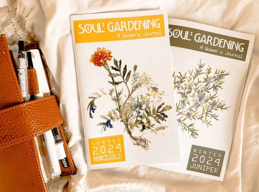 Two issues of Soul Gardening magazine, summer 2024 Marigold and winter 2024 Juniper, with illustrated botanical covers, resting on a white fabric surface, next to an orange leather pen case with pens.