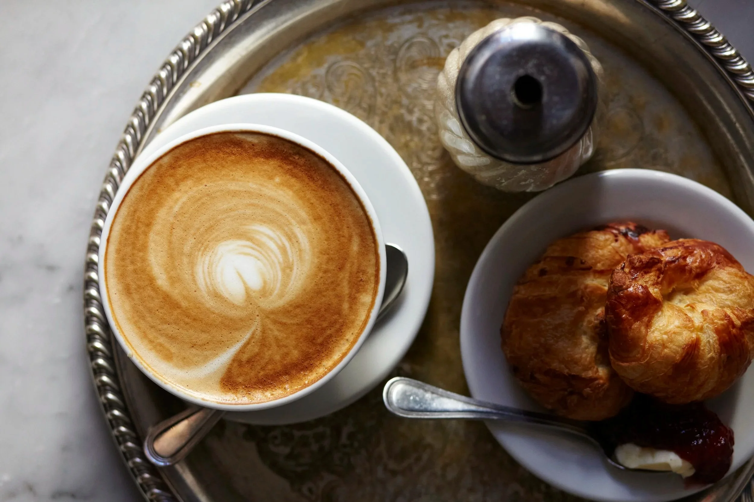 A cup of cappuccino with latte art, a croissant with jam and cream, and a bottle of sugar on a silver tray.