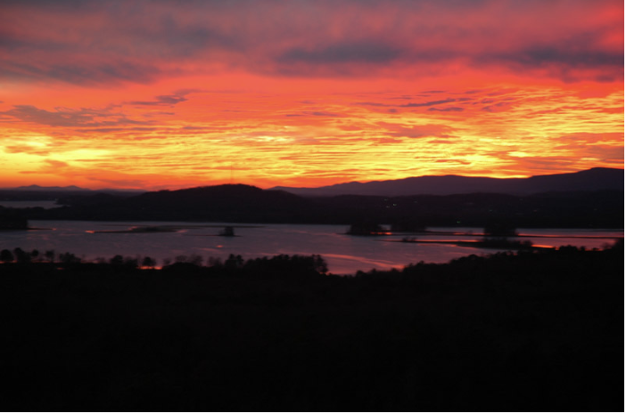 Vivid sunset over a body of water with distant hills and a dark foreground.