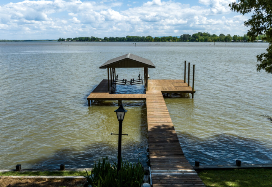 Wooden dock with a covered seating area extending into a calm lake, with a lamp post and greenery in the foreground, and trees and cloudy sky in the background.