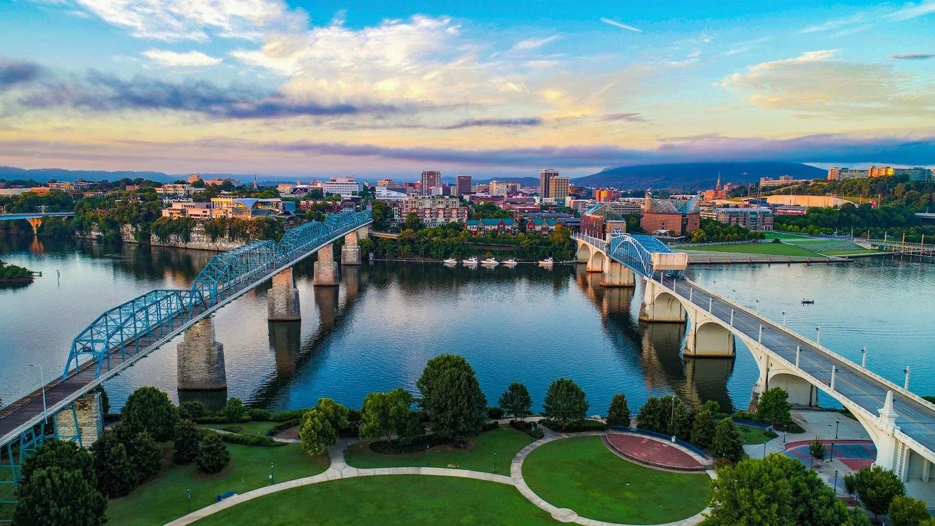 A cityscape view of downtown across a wide river, with bridges crossing over the river and a park with trees and walking paths in the foreground, under a partly cloudy sky.