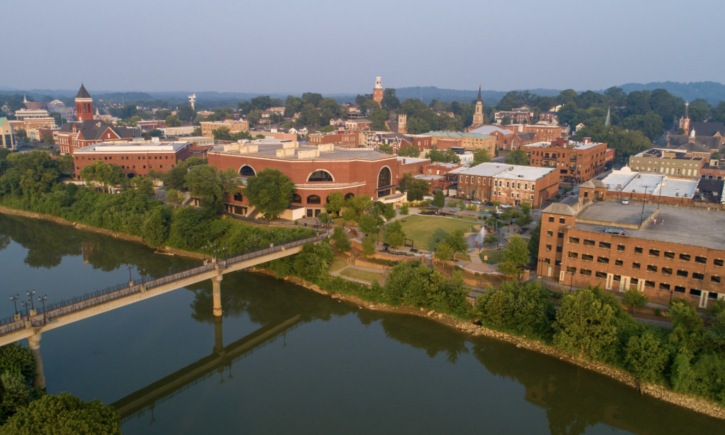 Aerial view of a downtown area with red-brick buildings, a river with a bridge, and green trees.
