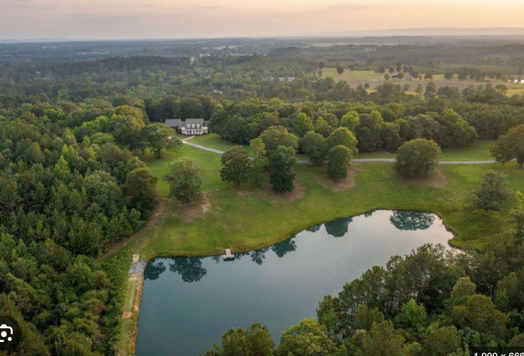 Aerial view of a large property with a pond, surrounded by trees and greenery, and a house in the background during sunset.