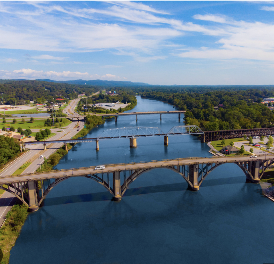 Aerial view of a river with multiple bridges crossing over it, surrounded by green trees and city roads, under a partly cloudy blue sky.