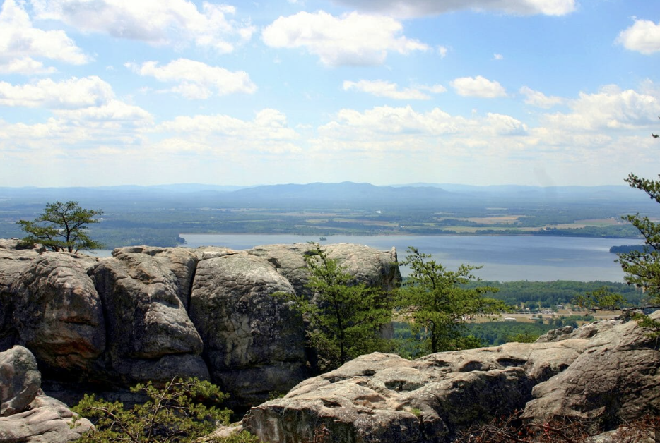 A scenic view of a lake and distant mountains seen from a rocky overlook with some green trees in the foreground and a partly cloudy sky.