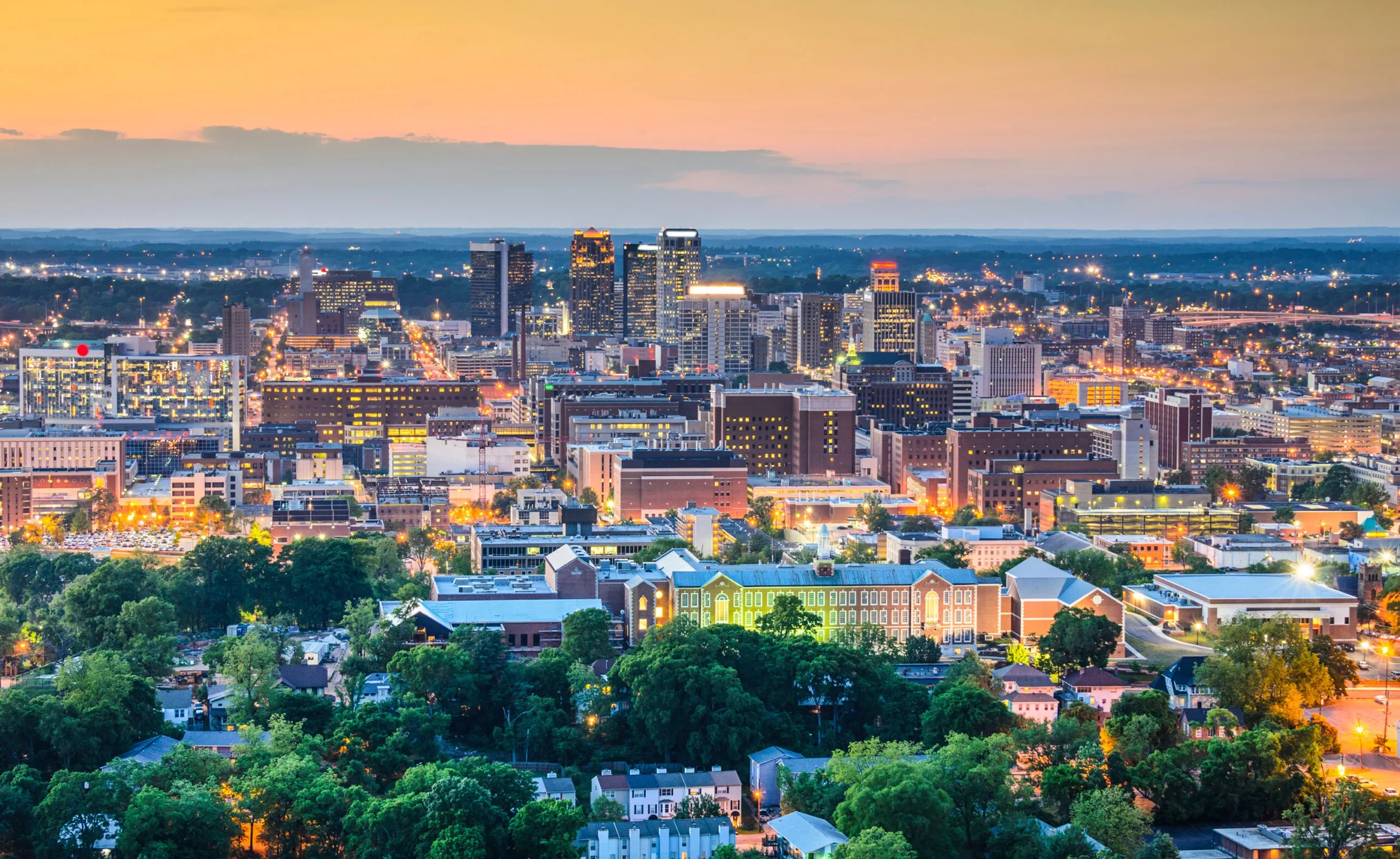 Aerial view of a city skyline at dusk with illuminated buildings and a large green park in the foreground.