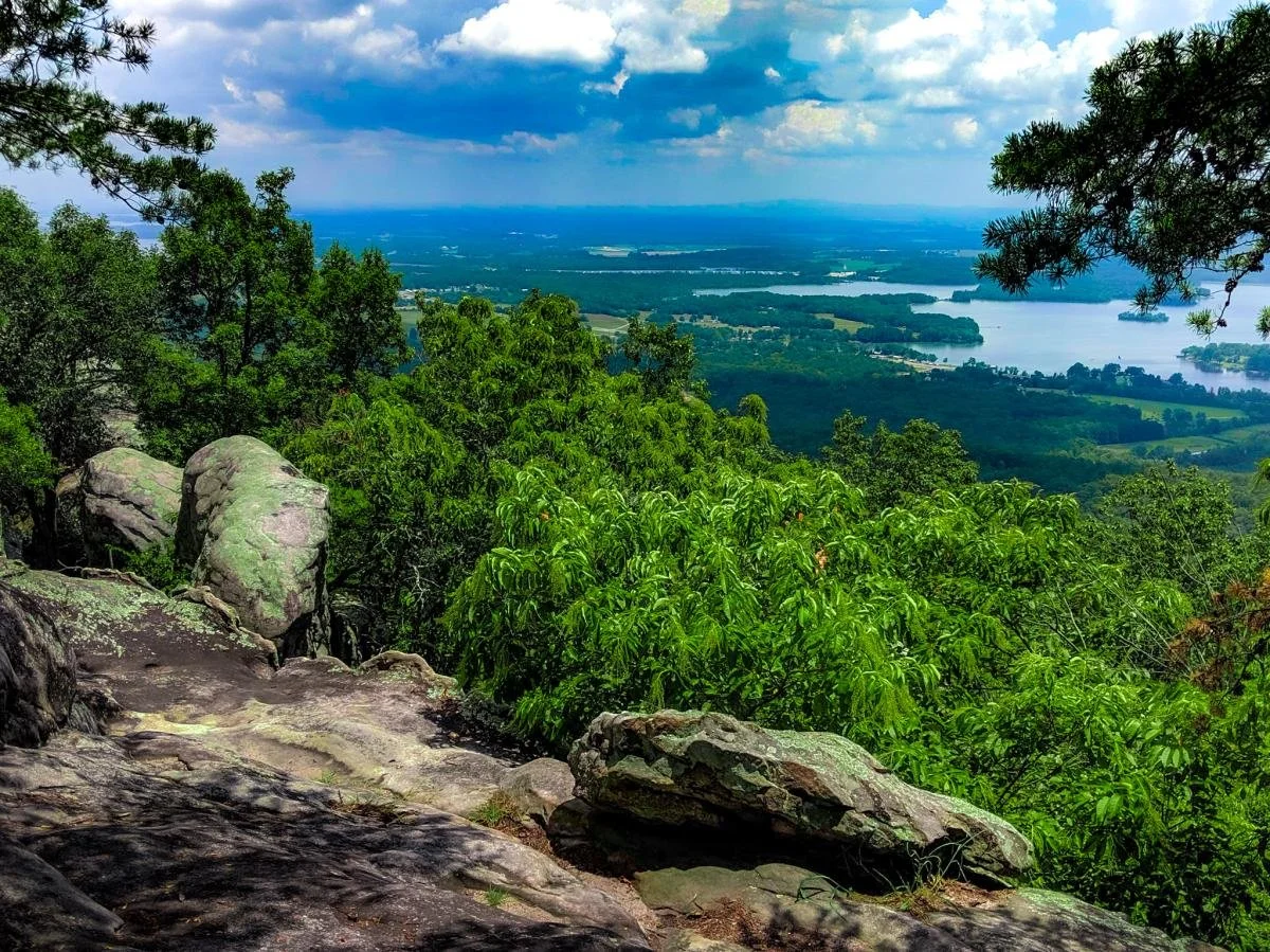 View from a mountain with rocks and green trees overlooking a landscape of lakes and fields under a partly cloudy sky.