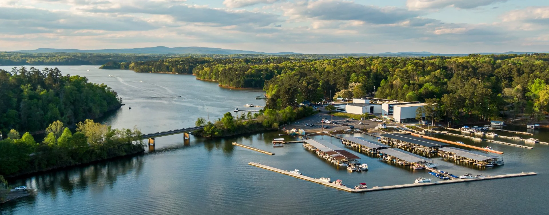 A scenic view of a marina with floating docks and boats, surrounded by lush green trees, with a river and bridge in the background, and distant hills under a partly cloudy sky.