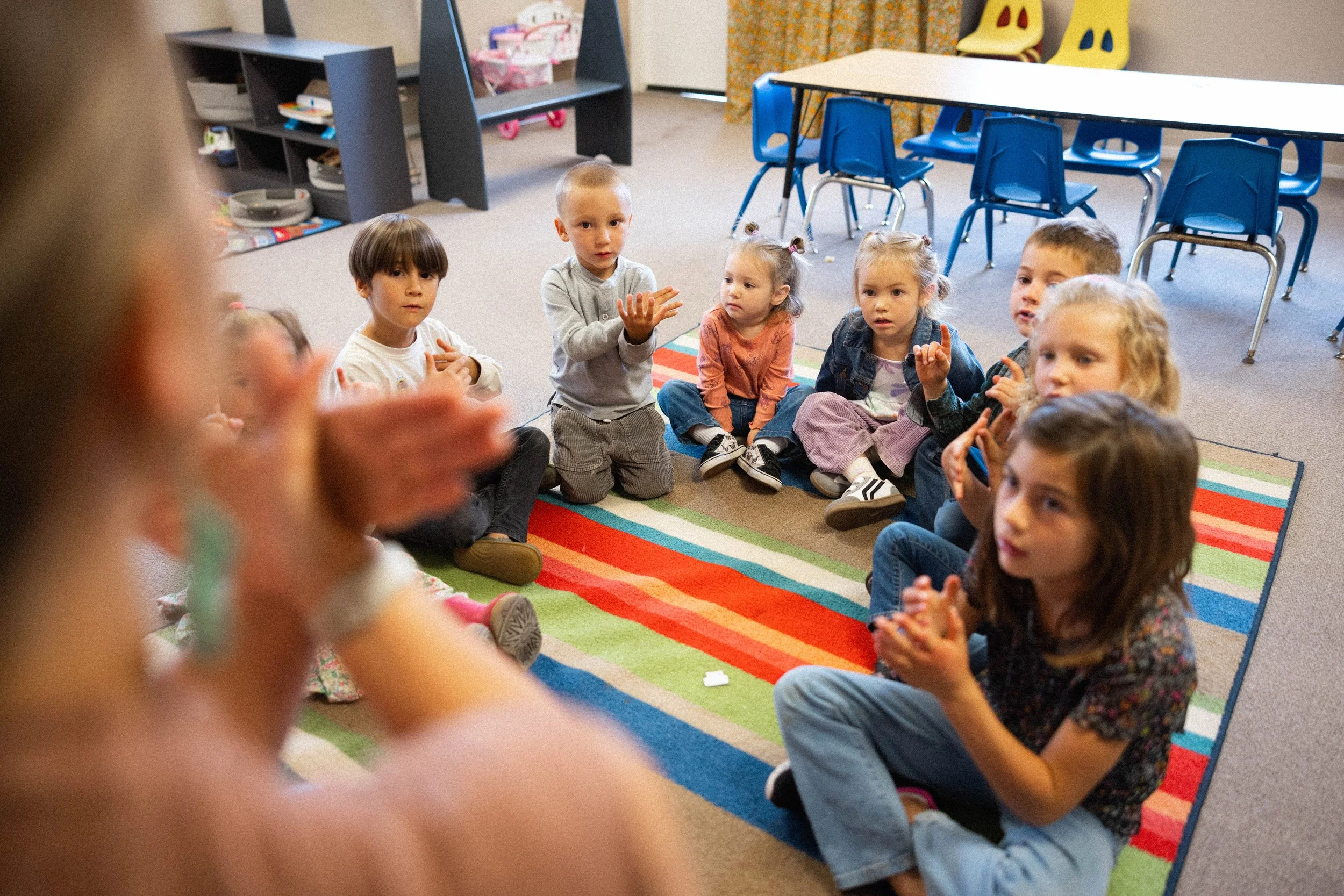 A group of young children sitting in a semi-circle on a colorful striped rug in a classroom, engaged in a discussion with an adult, with some looking attentive and others gesturing with their hands.