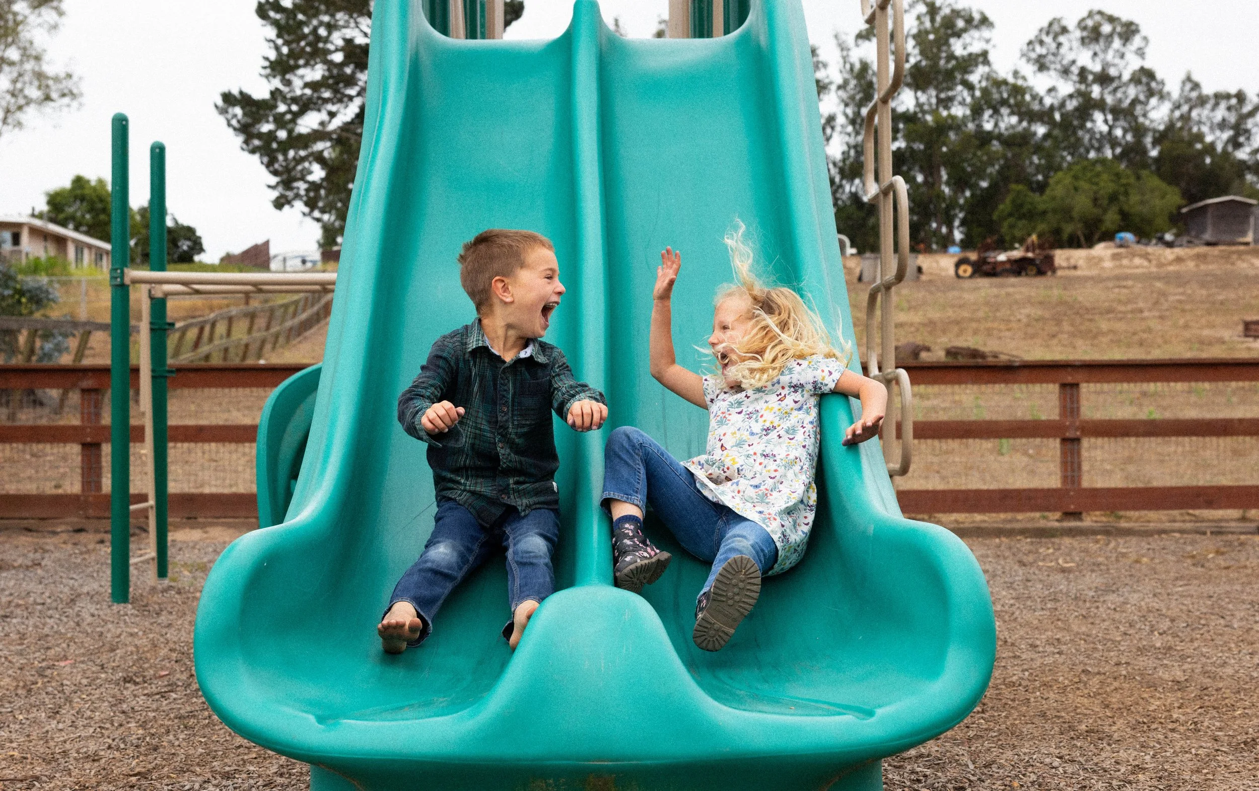 Two children, a boy and a girl, are playing and laughing together on a blue slide at a playground. The boy is sitting at the top of the slide, while the girl is sitting lower down, both expressing joy and excitement.