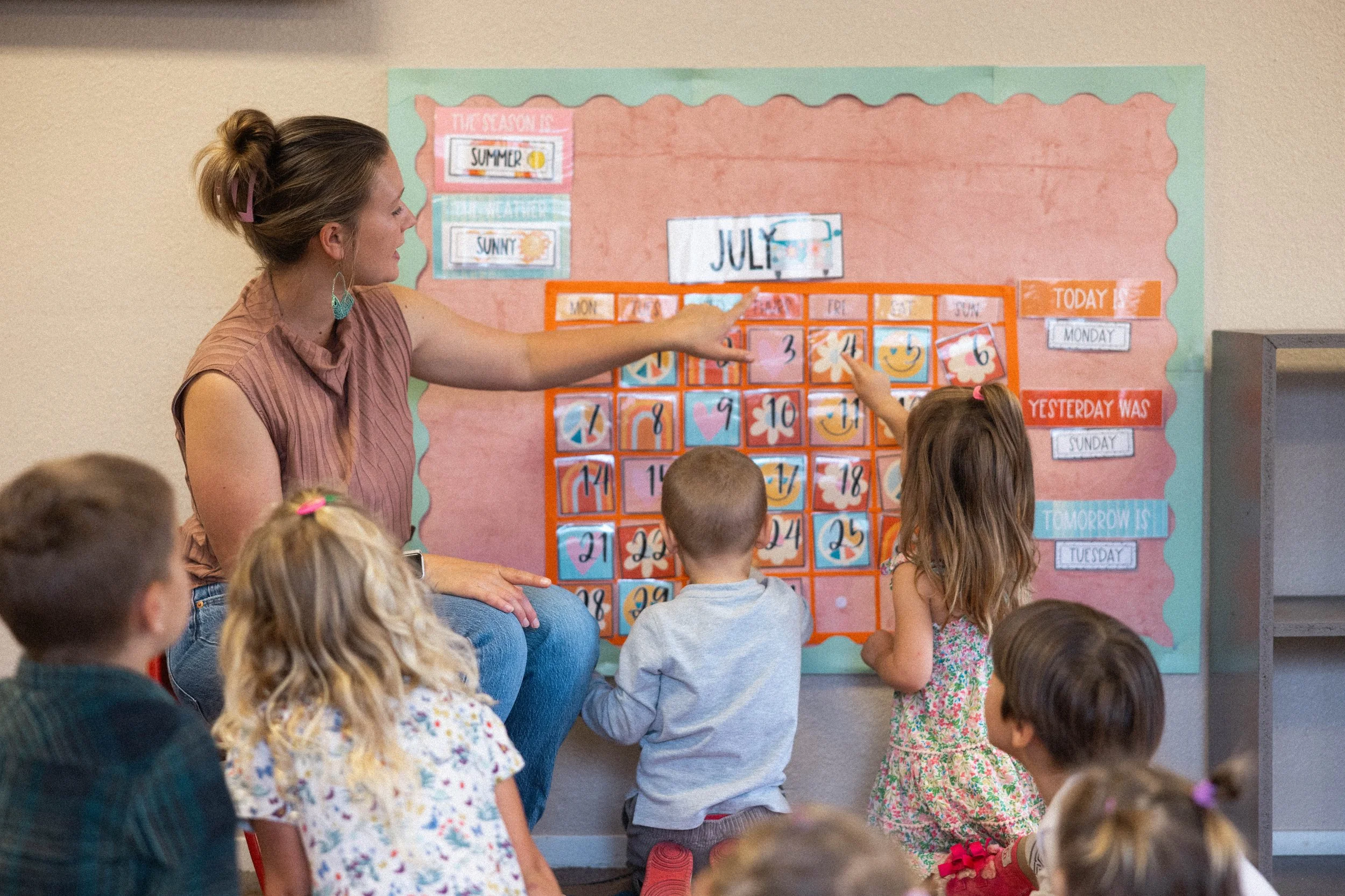 A teacher sitting on a chair points to a colorful classroom calendar on the wall, surrounded by young children sitting on the floor, learning about the month of July.