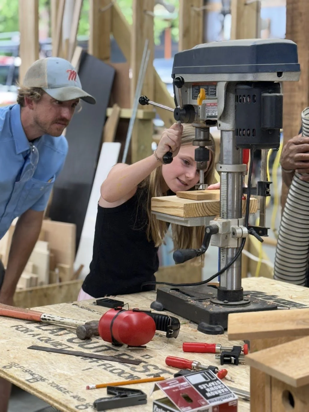 A girl operating a drill press while an adult watches in a woodworking shop.