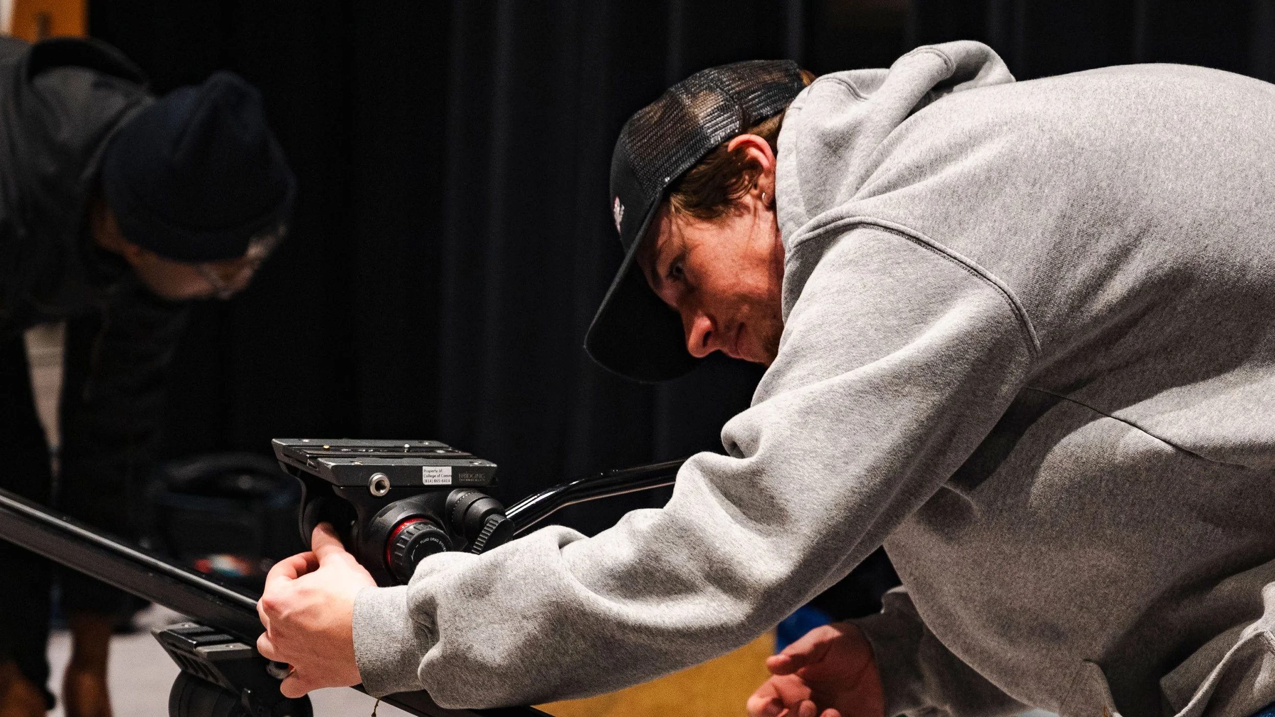 A man in a gray hoodie and black cap is operating a camera on a skateboard, with another person in a black hoodie and glasses in the background.