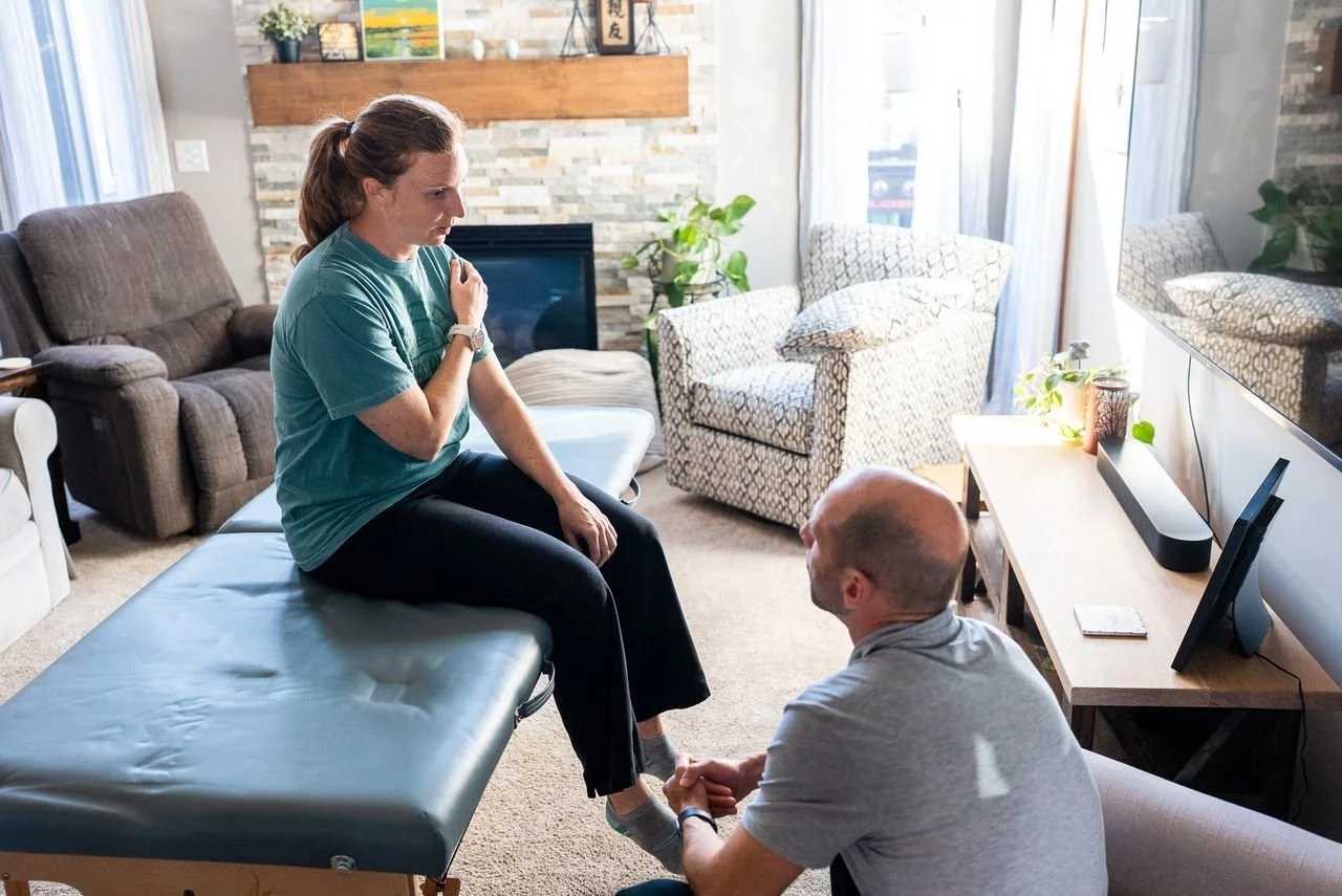 A woman sitting on an examination table with her hand on her shoulder, talking to a man who is kneeling in front of her, inside a living room with a TV, sofa, and armchair.