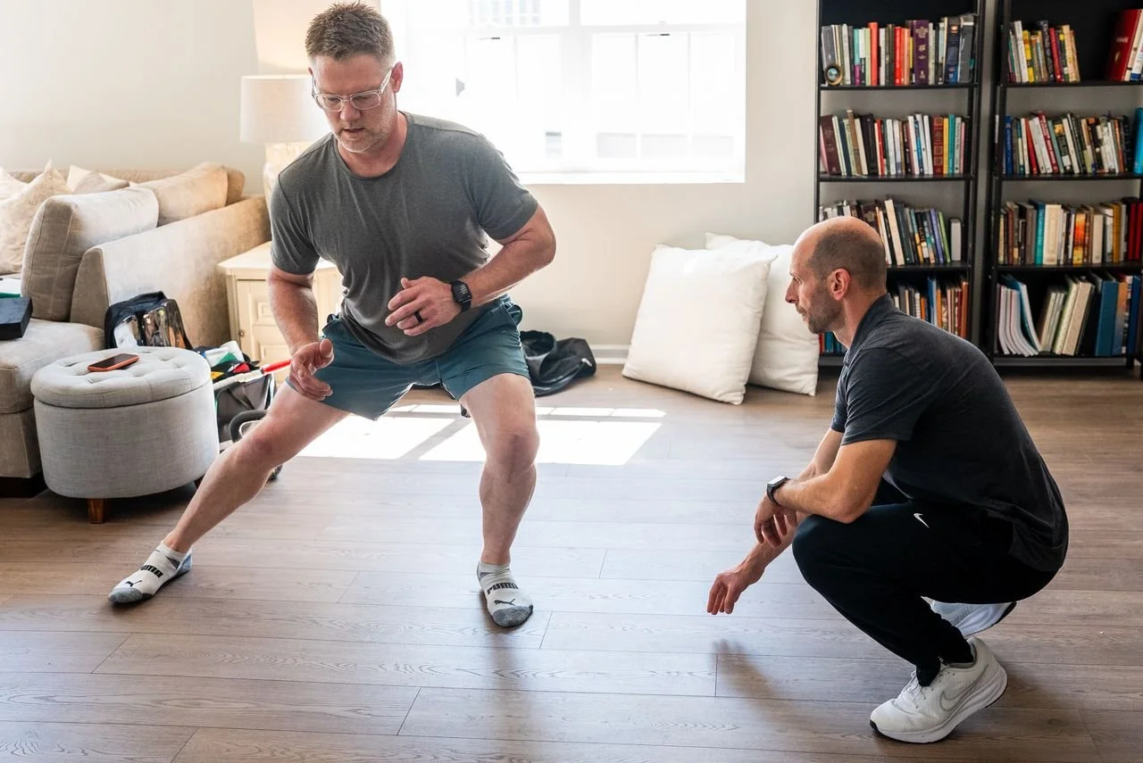 Two men exercising indoors, one is in a squat position while the other is crouching and observing, in a living room with bookshelves and a sofa.
