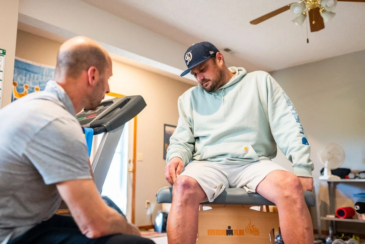 A man sitting on an examination table with his eyes closed, wearing a hoodie and shorts, while another man kneels beside him. The room has a ceiling fan, a fan on a table, and various fitness equipment.