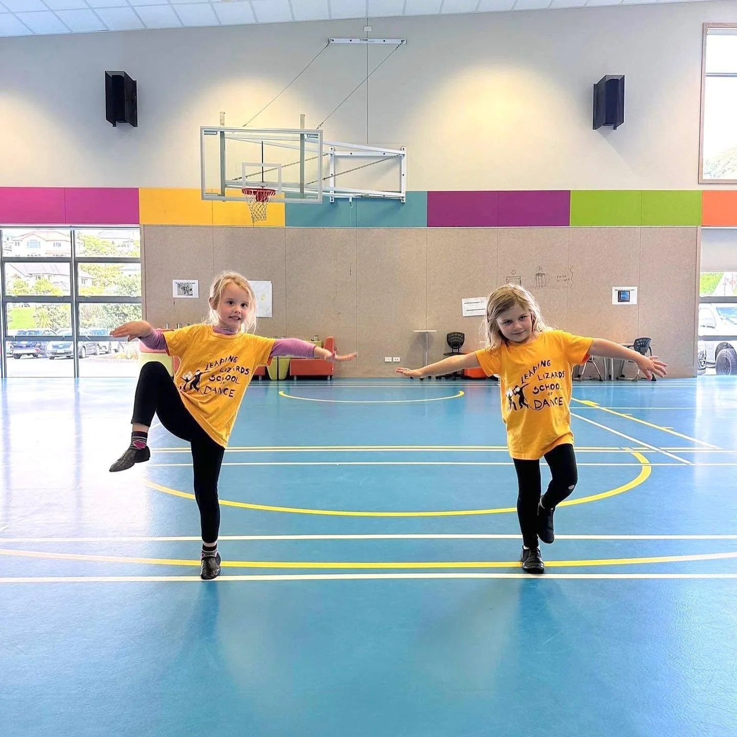 Two young girls in yellow T-shirts performing a dance pose in an indoor gymnasium with colorful wall panels and large windows in the background.