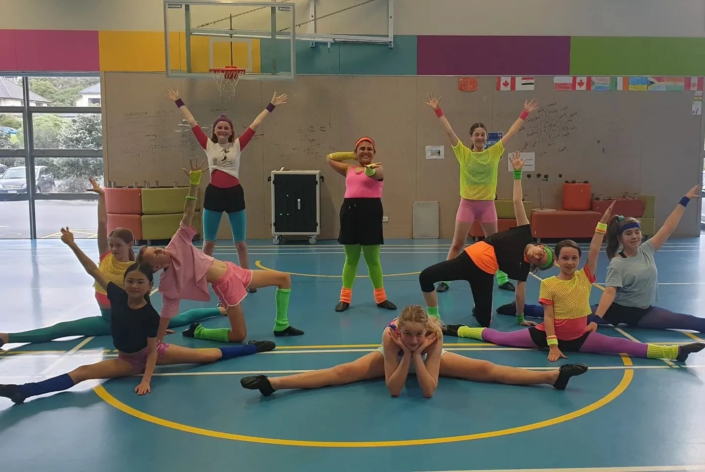 Group of children and teens in colorful dance clothes posing in a gymnasium with a blue floor and large windows.