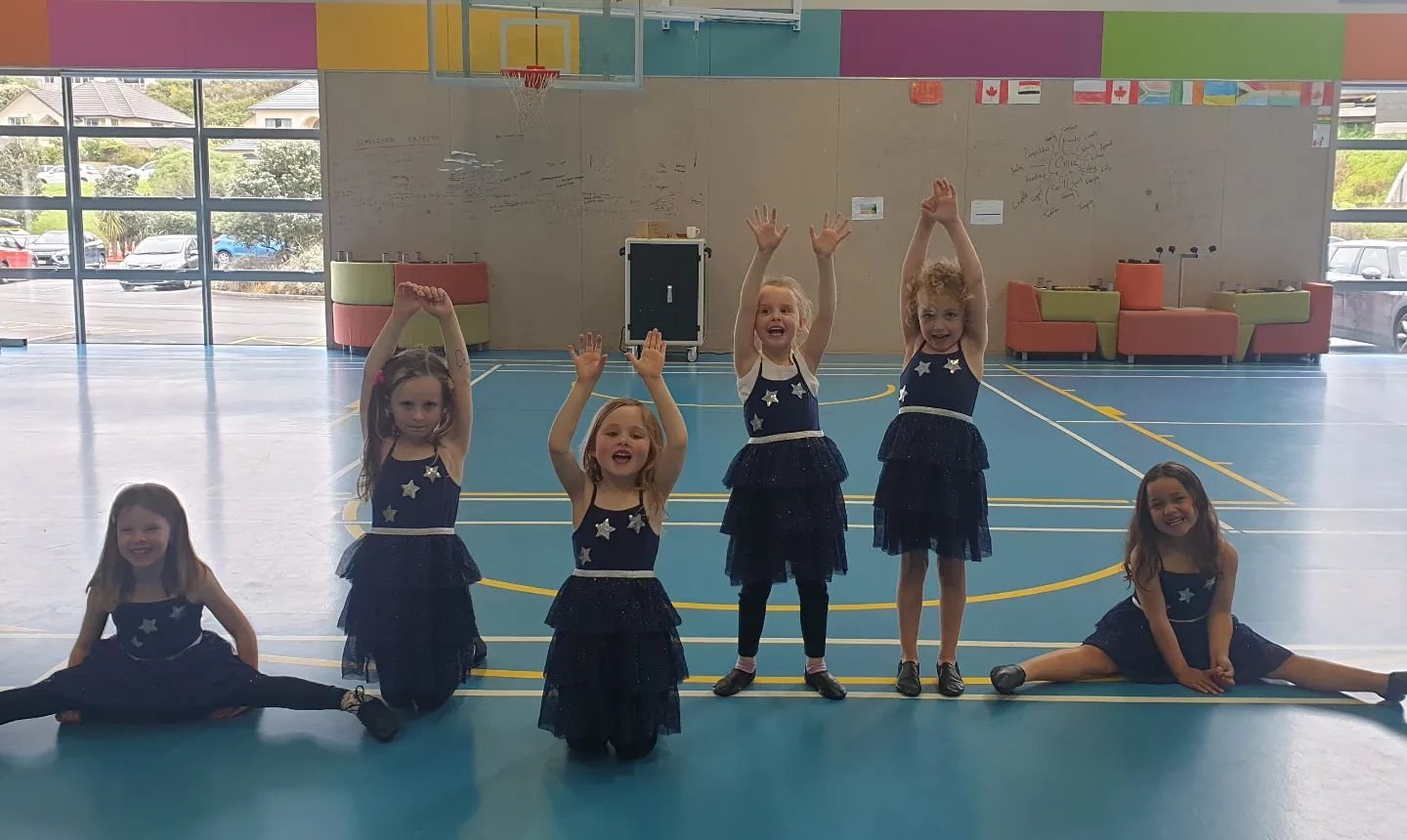 Six young girls in matching dark blue dresses with star patterns and ruffled skirts pose in a gymnasium, some sitting or doing splits on the floor and others standing with arms raised..