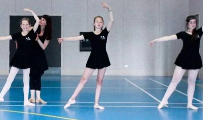 Three young girls in black dance leotards and skirts practicing ballet in a gymnasium, while a female teacher corrects them.