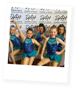 Group of 5 girls in blue and green sparkly dance costumes, posing for the camera in front of a sign saying "DAH dance-a-holic"