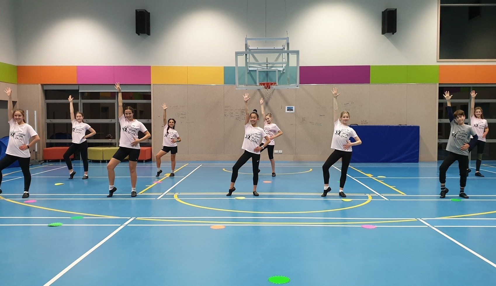 Group of young dancers practicing in a gymnasium with colorful wall accents and spot markers on the floor.