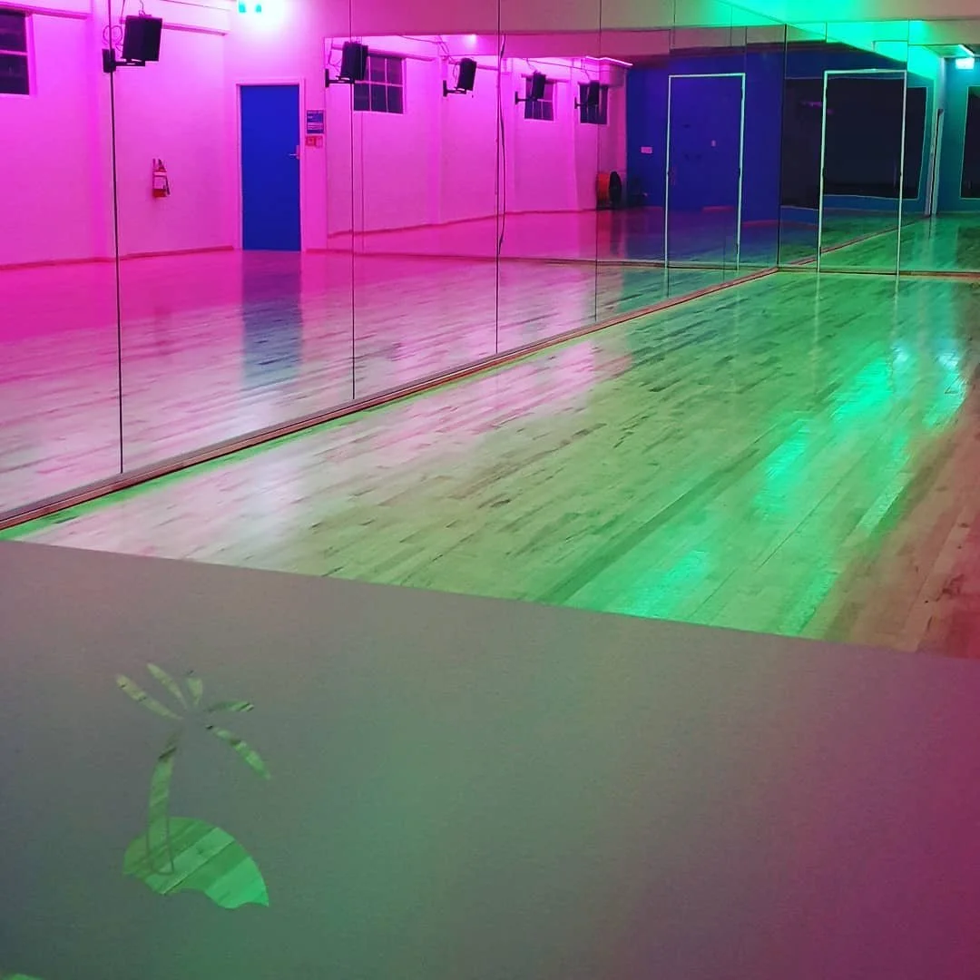 An empty dance studio with wooden floors, mirrored wall, and colorful ambient lighting.