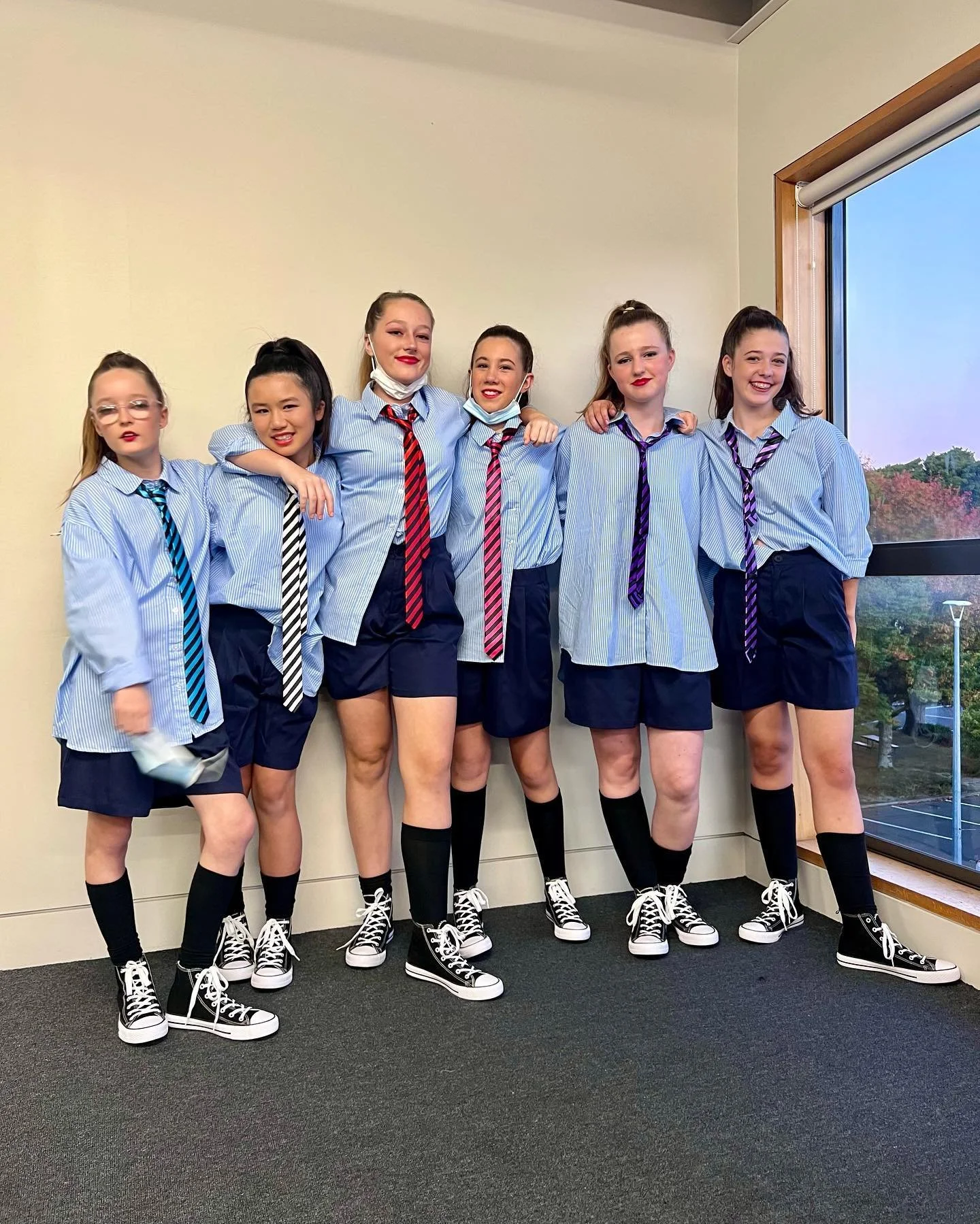 Group of six girls in blue and white school uniforms with striped ties, standing together in a room near a window, smiling.