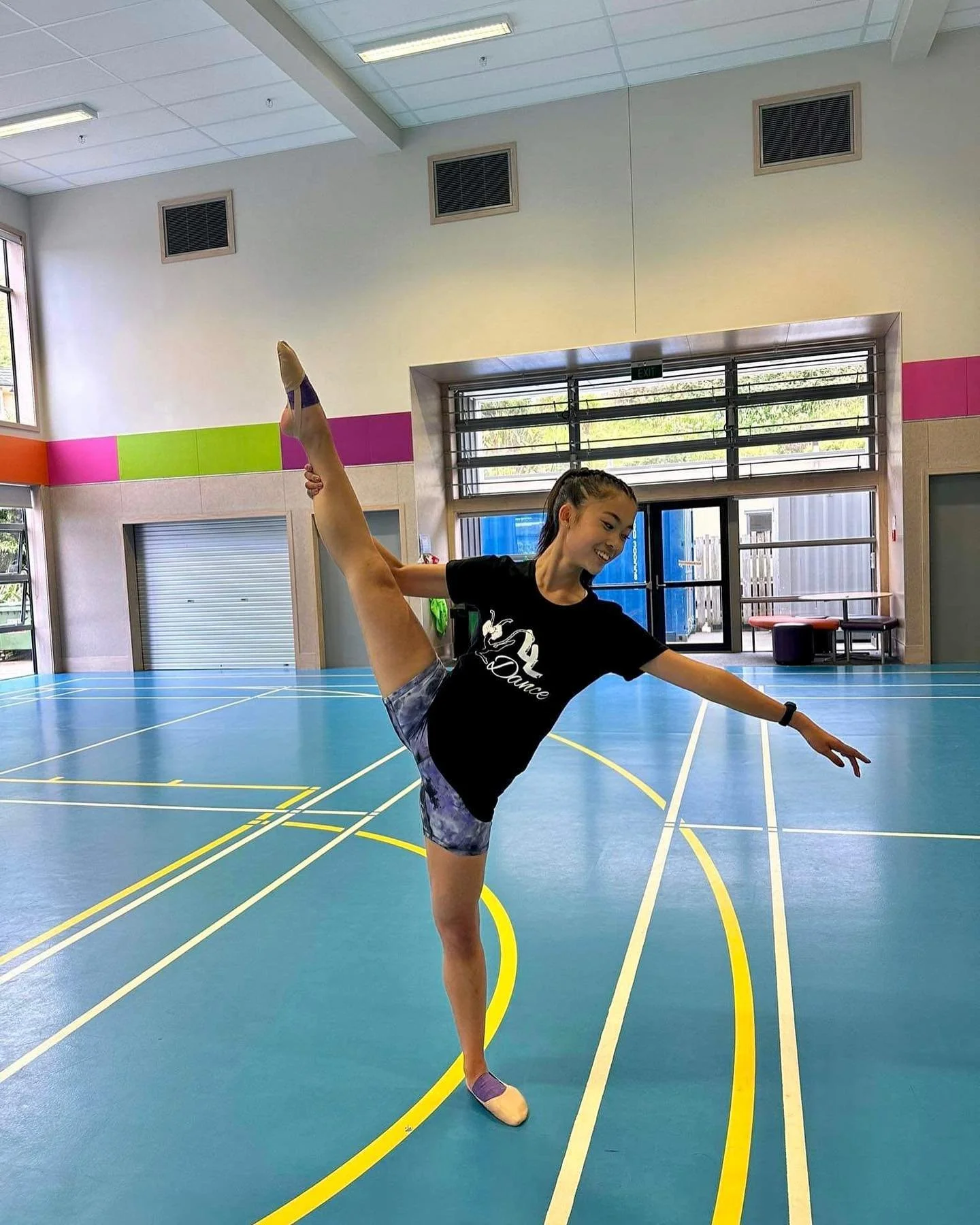 A young woman practicing dance in a gymnasium, standing on one foot while holding her other leg extended to the side, above her head. She is smiling and wearing a black t-shirt, shorts, and ballet slippers.