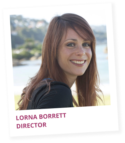 A woman with long red hair smiling outdoors near water, wearing a black top, with a scenic background.