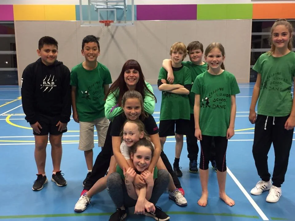 Group of children and a dance teacher posing in a gymnasium, some children wearing green shirts with text, and a few standing with arms crossed or smiling.