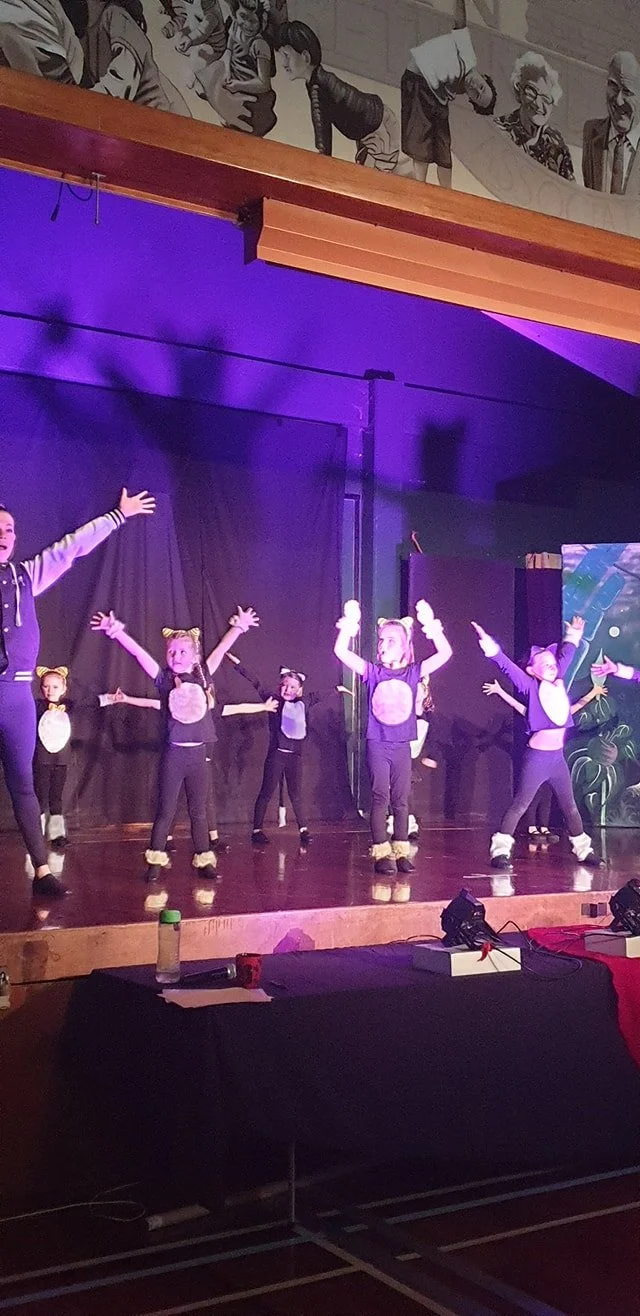Children performing a dance on stage dressed as mice, with some wearing mouse ears and costumes, under purple stage lighting.