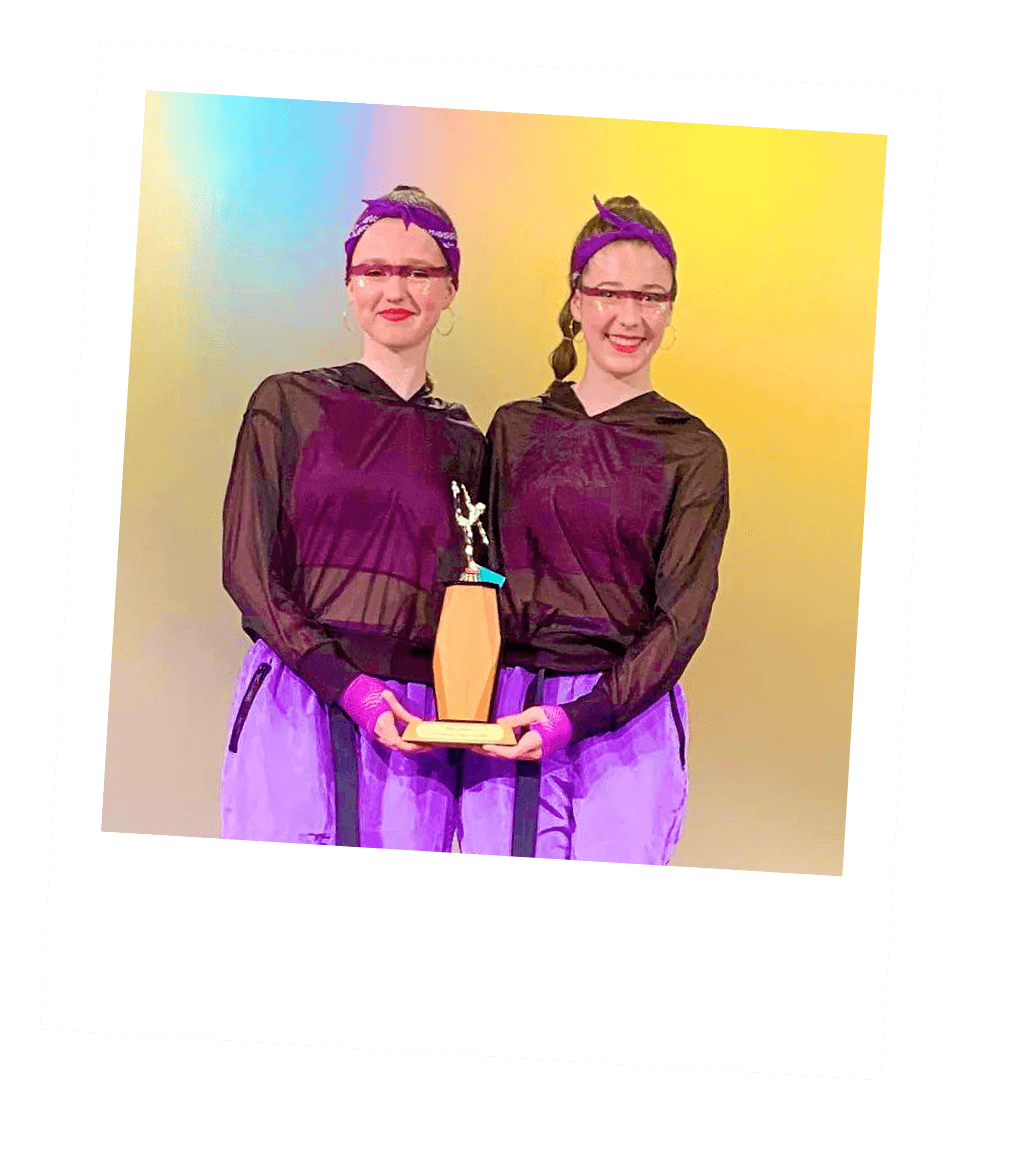 Two women in matching outfits holding a trophy, smiling, standing against a yellow background.