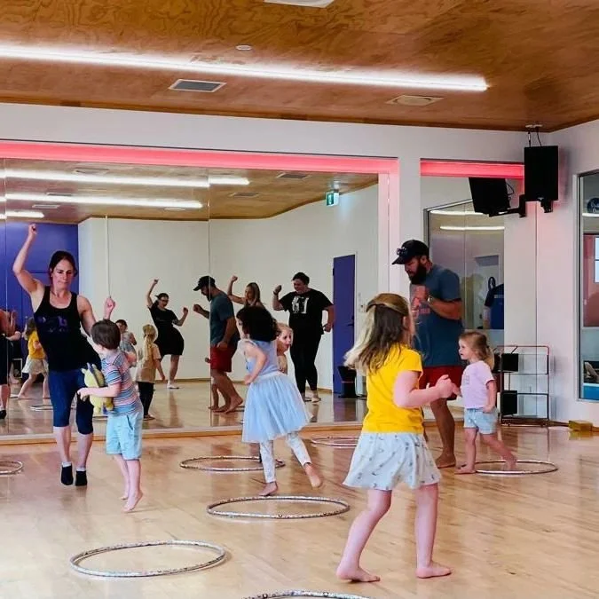 Children and adults participating in a dance class in a studio with a large mirror, wooden floors, and colorful lighting.