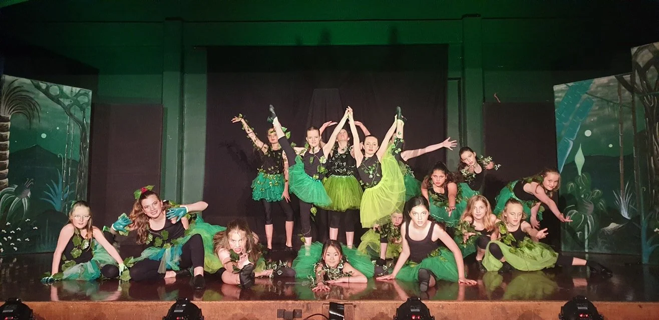 Group of children performing a theatrical dance on stage, dressed in black and green costumes with a nature theme.