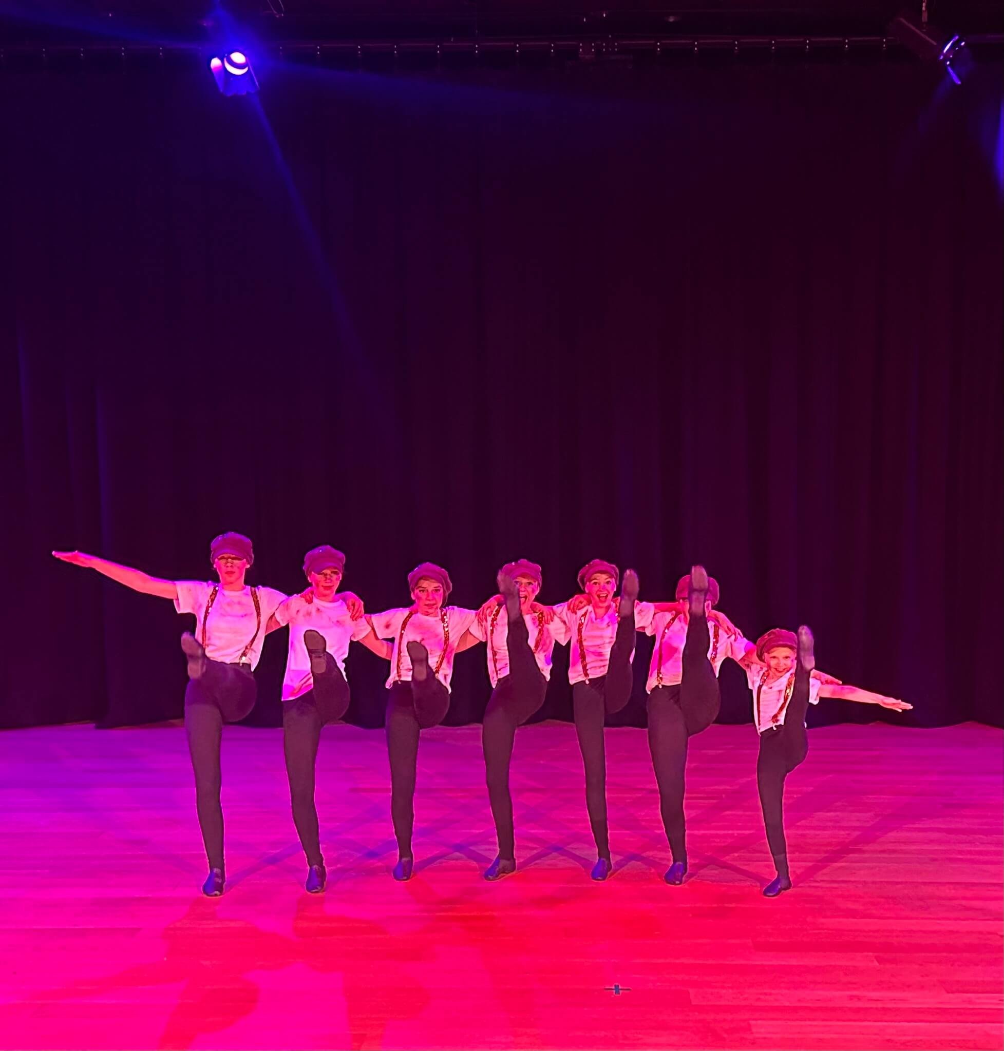 A group of seven dancers in black tights, white shirts, and suspenders performing on a stage with dark curtains, illuminated by colorful stage lights.